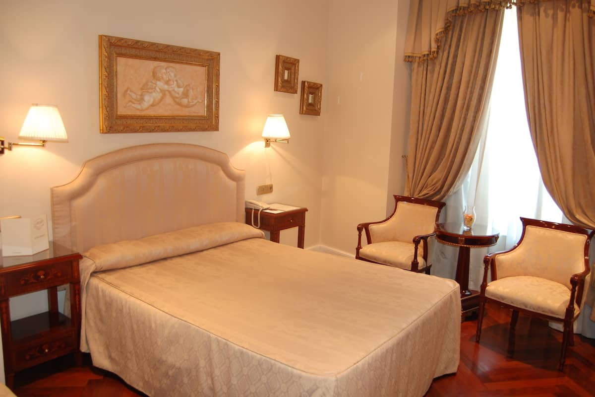 Une chambre d’hôtel avec un grand lit, deux chaises, une table de chevet, une lampe et un tableau encadré au mur.