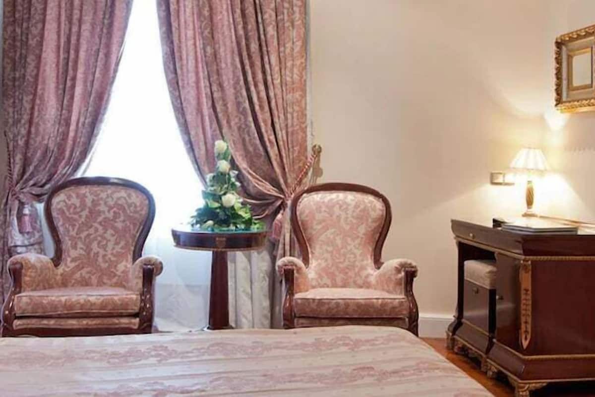 Une chambre d’hôtel comprenant un lit, deux fauteuils, une table d’appoint avec une lampe, une petite table avec un vase de fleurs et une commode avec un miroir.