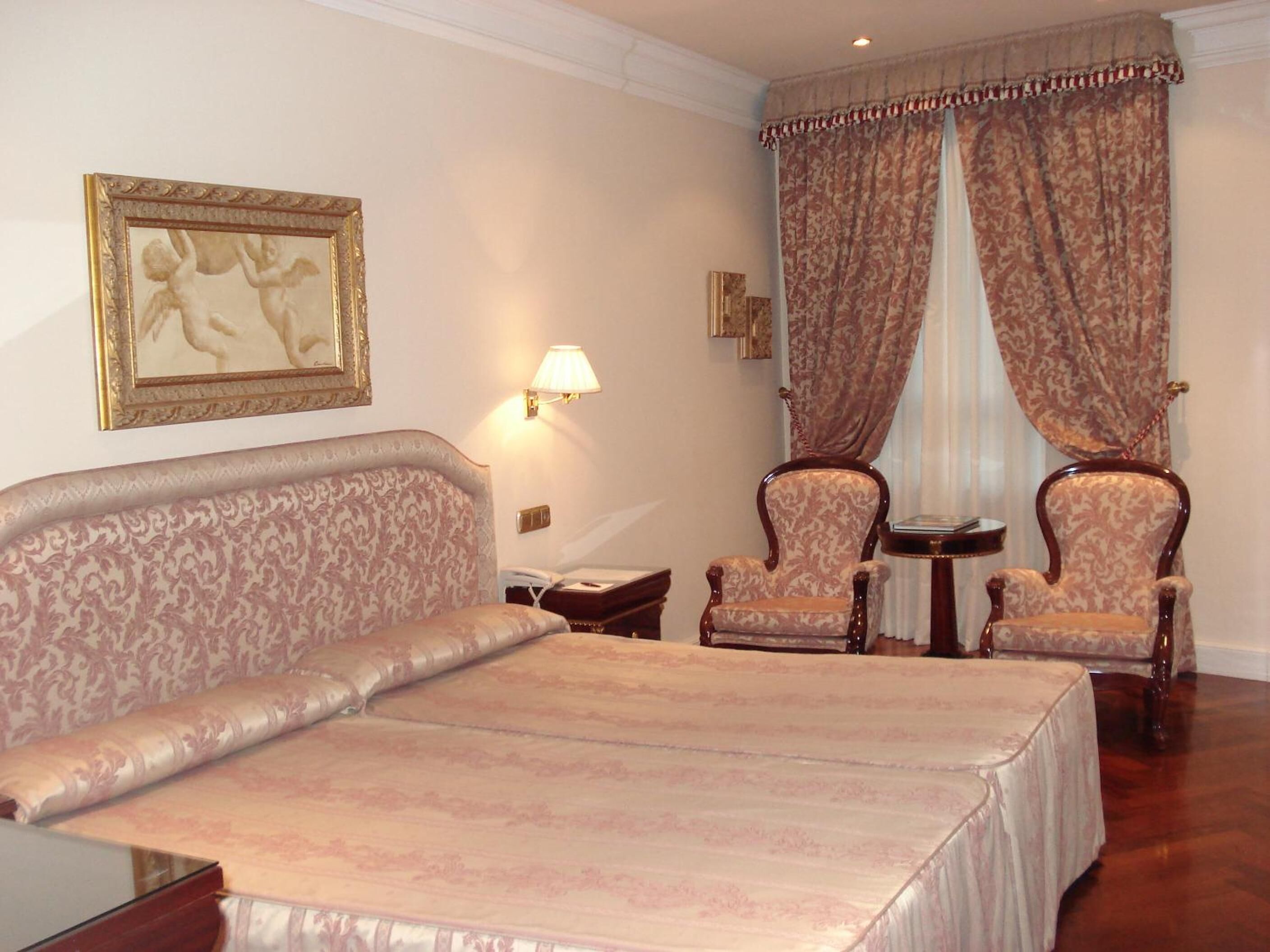 Una habitación de hotel con una cama grande, dos sillones, una mesita pequeña y una lámpara de pared.