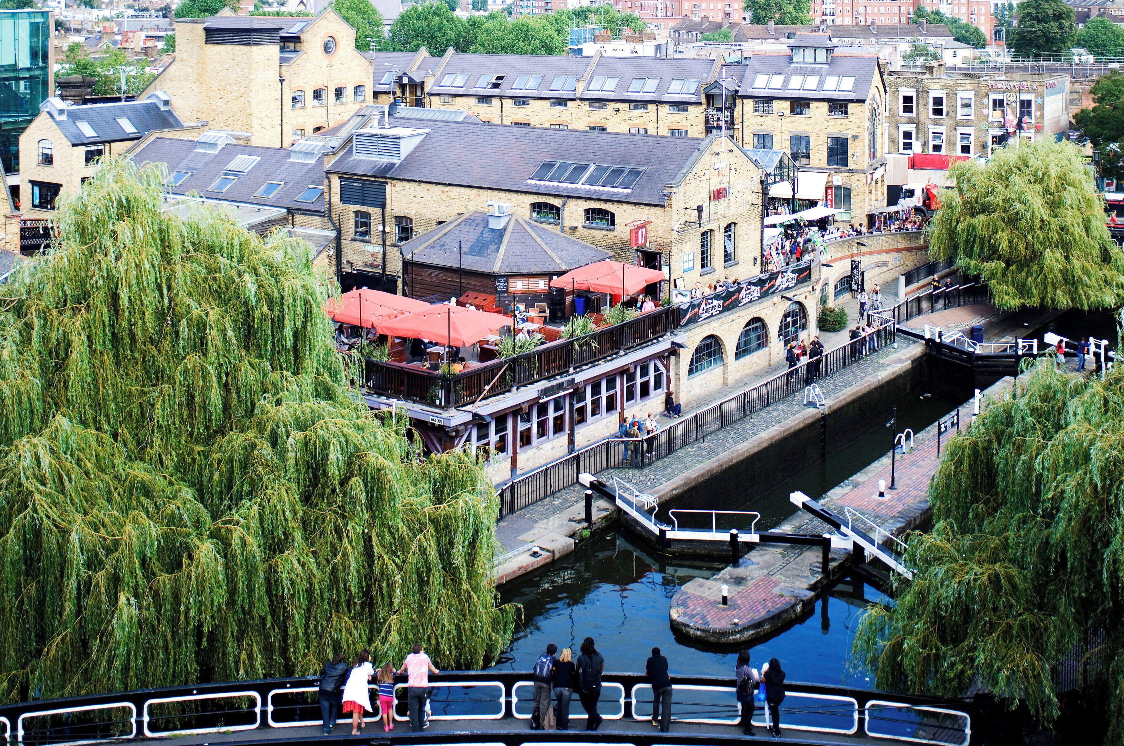A riverside walkway with a bridge connecting buildings, a canal, and willow trees.