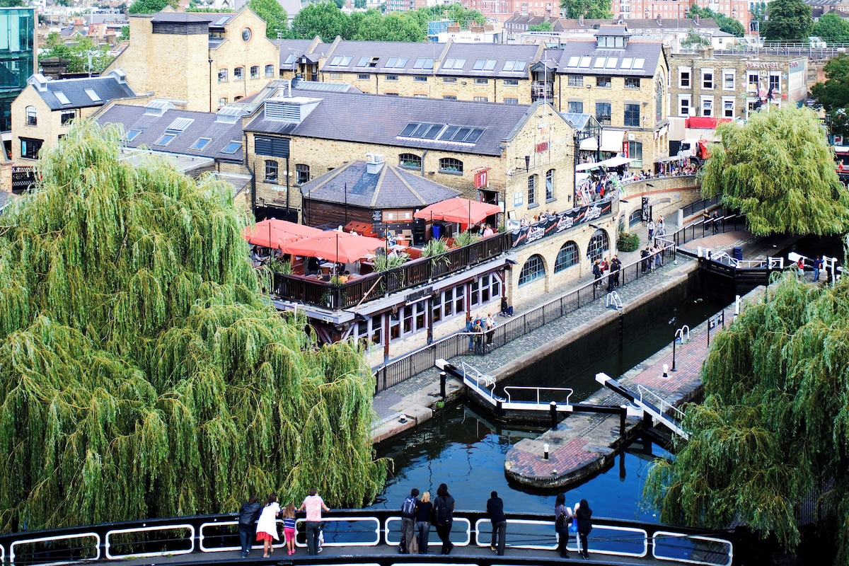 A riverside walkway with a bridge connecting buildings, a canal, and willow trees.