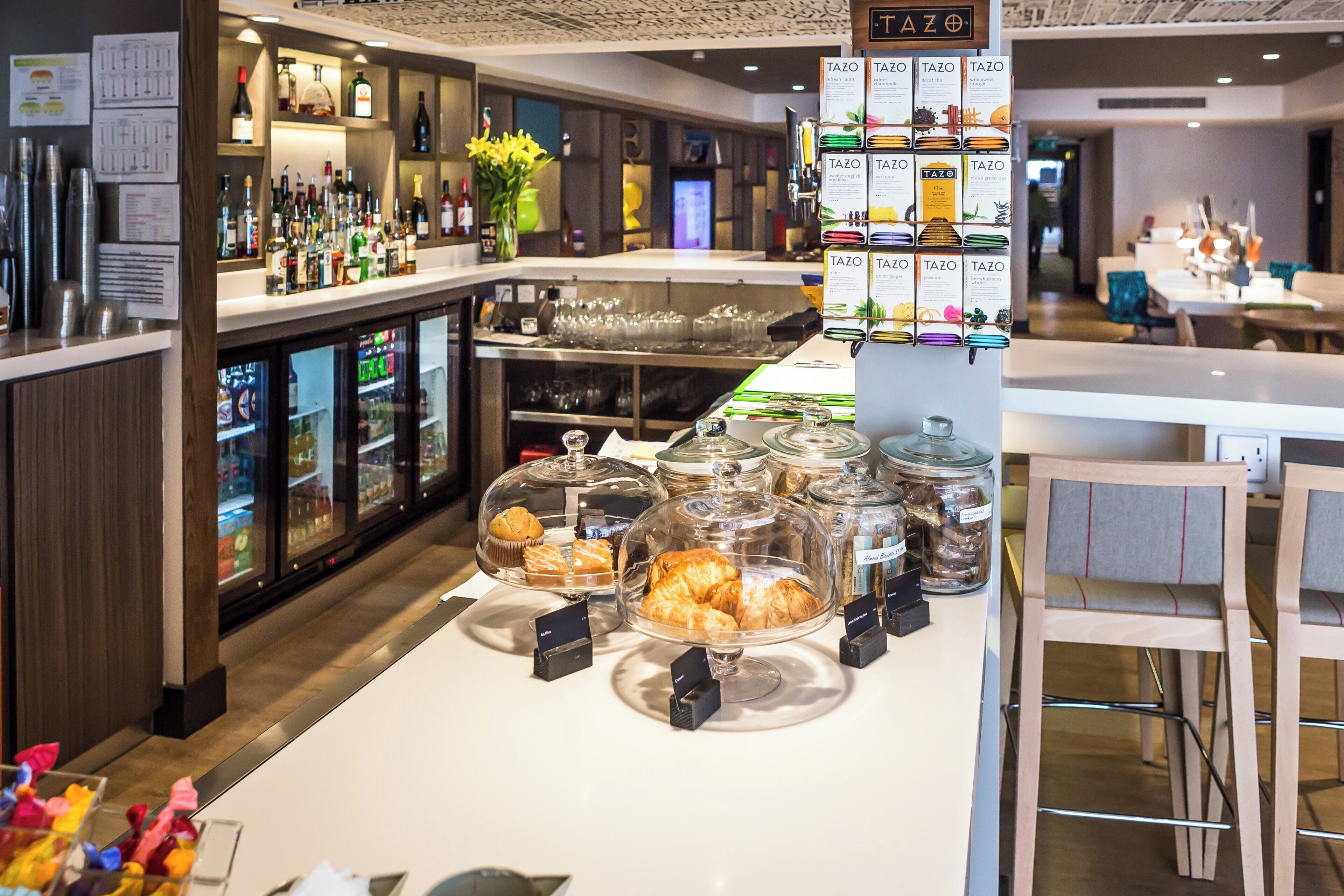 A hotel bar area with a variety of beverages, a display of TAZO tea boxes, and a dessert counter with pastries.