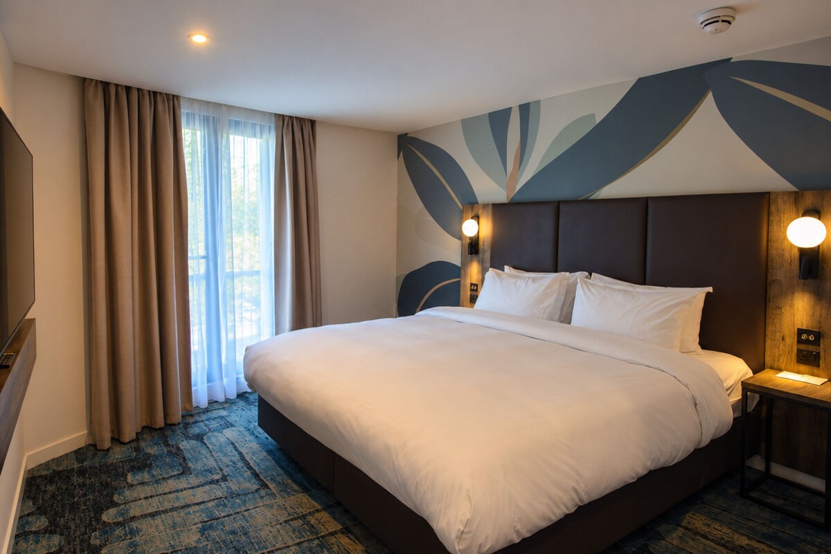 A hotel room with a large bed, a flat-screen TV, a desk with a lamp, and a view of the outdoors through the window.