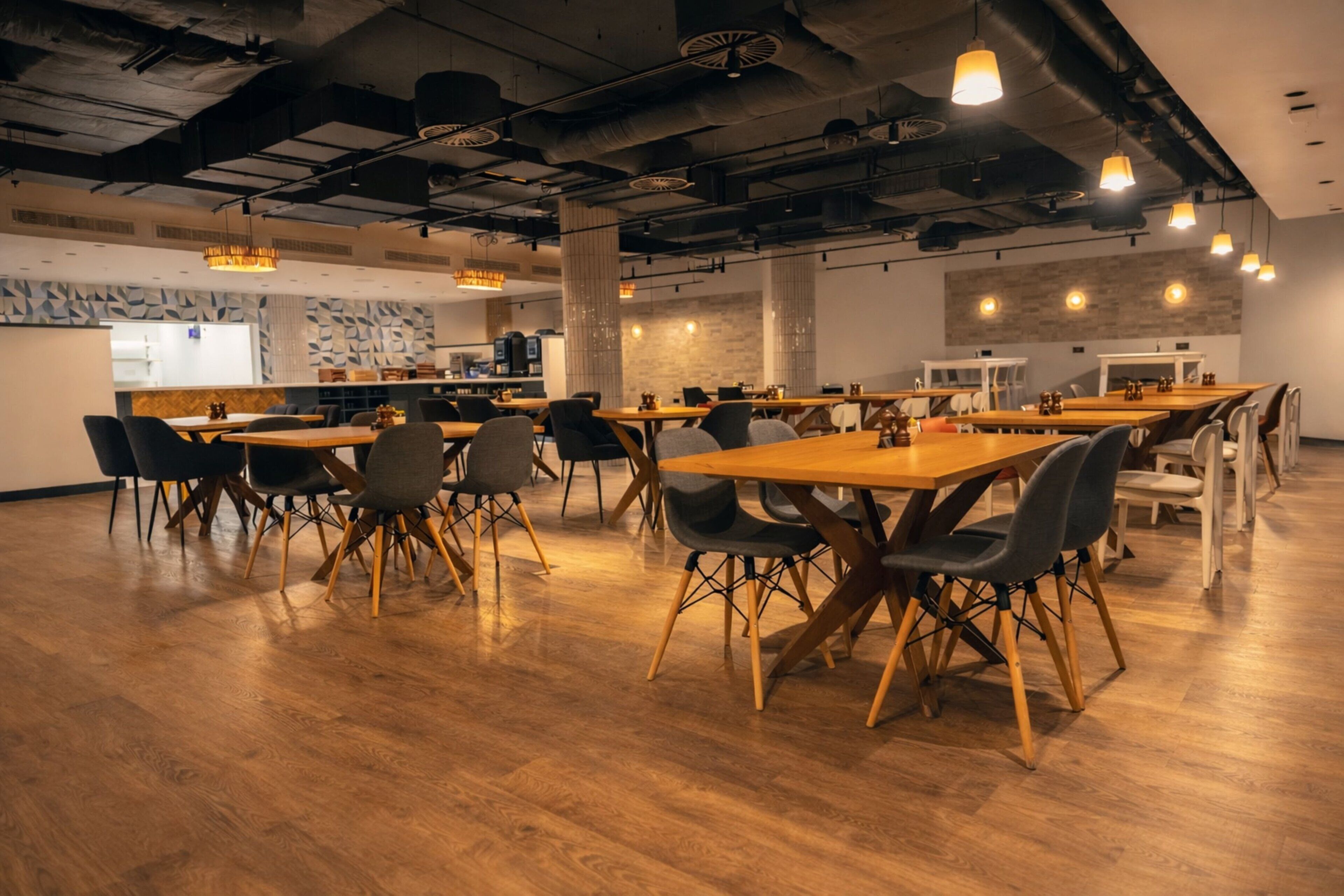 A spacious dining area with wooden tables and chairs, a kitchen in the background, and exposed ductwork on the ceiling.