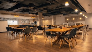 A spacious dining area with wooden tables and chairs, a kitchen in the background, and exposed ductwork on the ceiling.