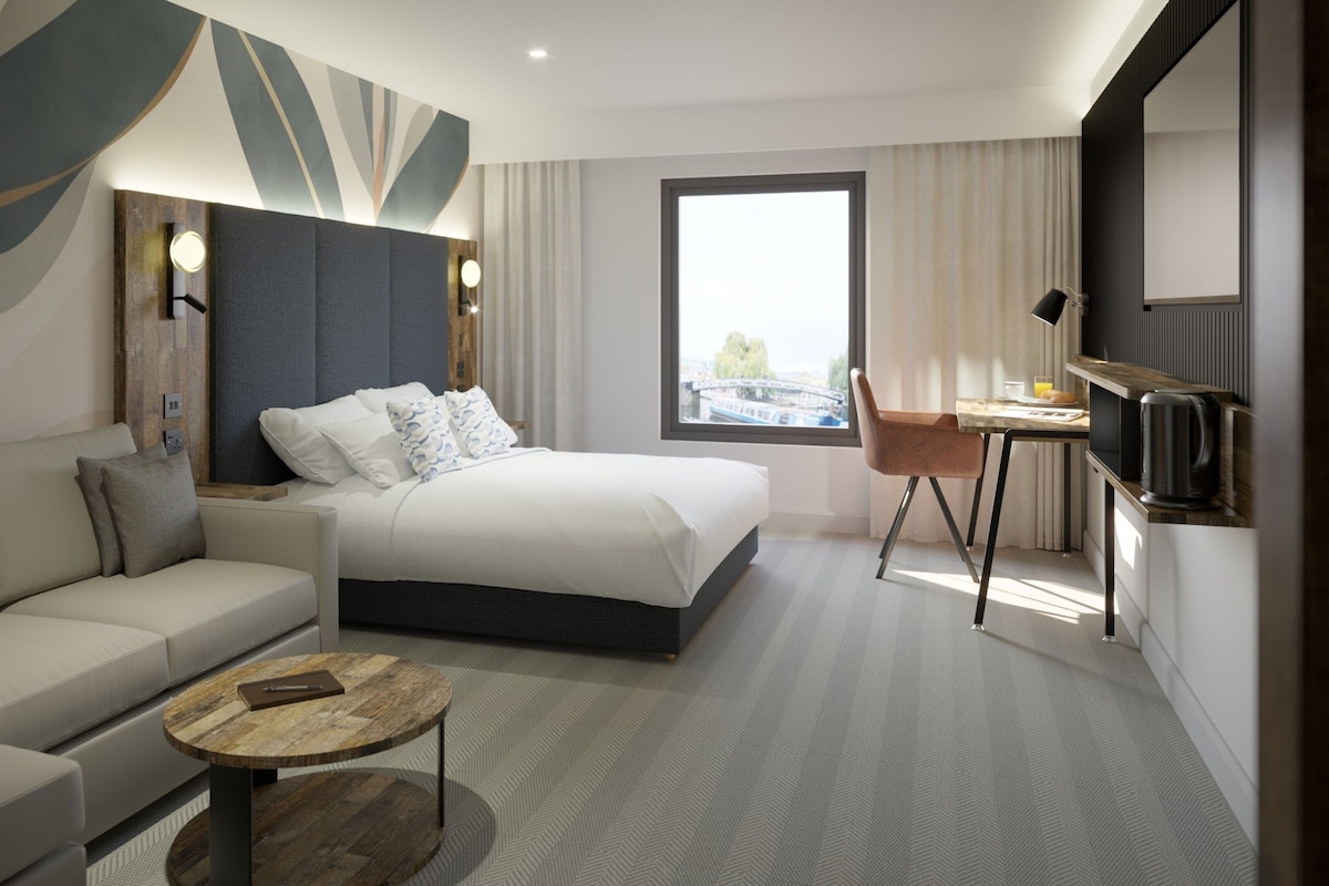 A modern hotel room with a bed, sofa, coffee table, and a dining area with a chair and table.