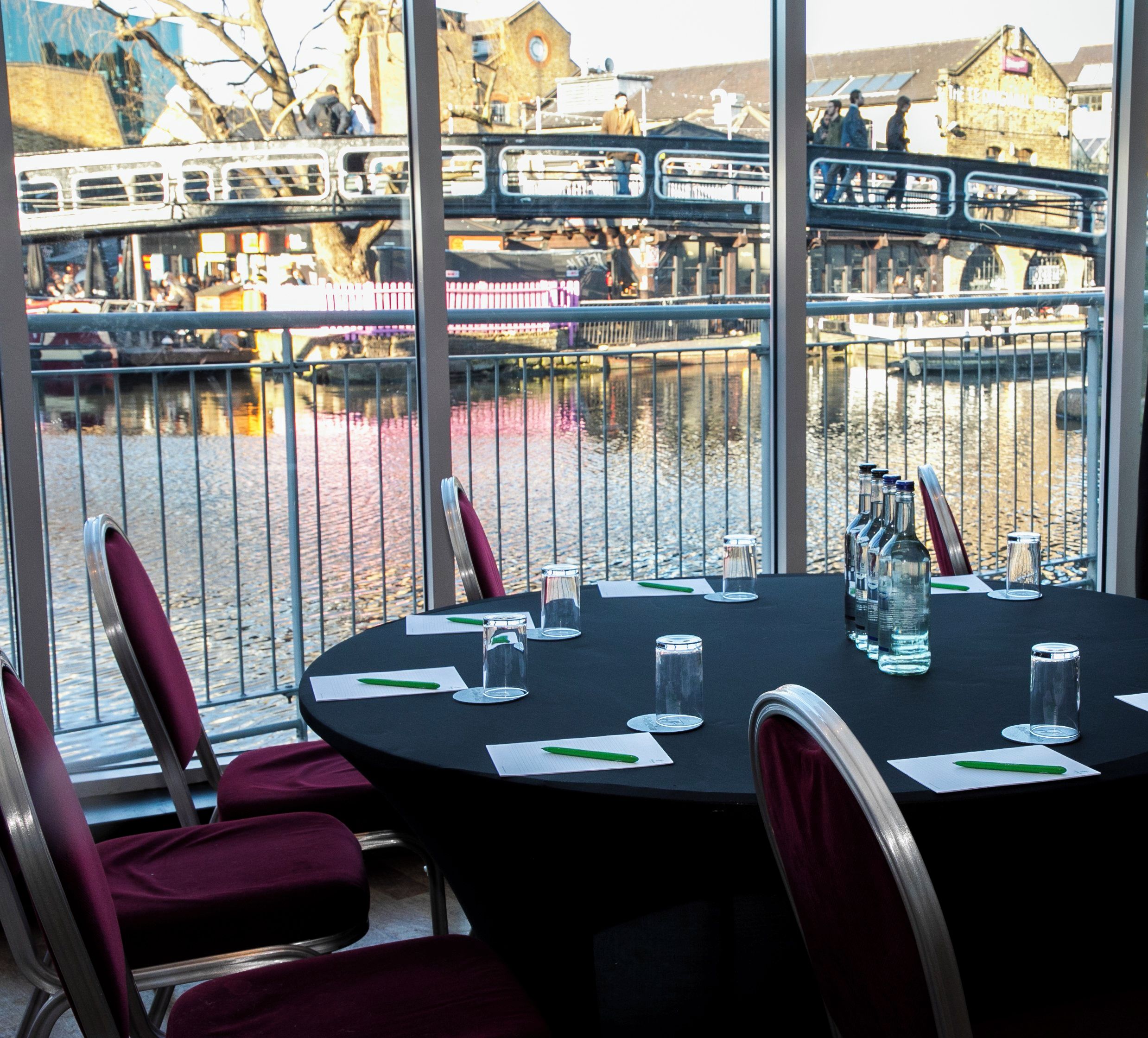 Un comedor interior con vista a un canal y edificios. Hay mesas dispuestas con platos, vasos y cubiertos.