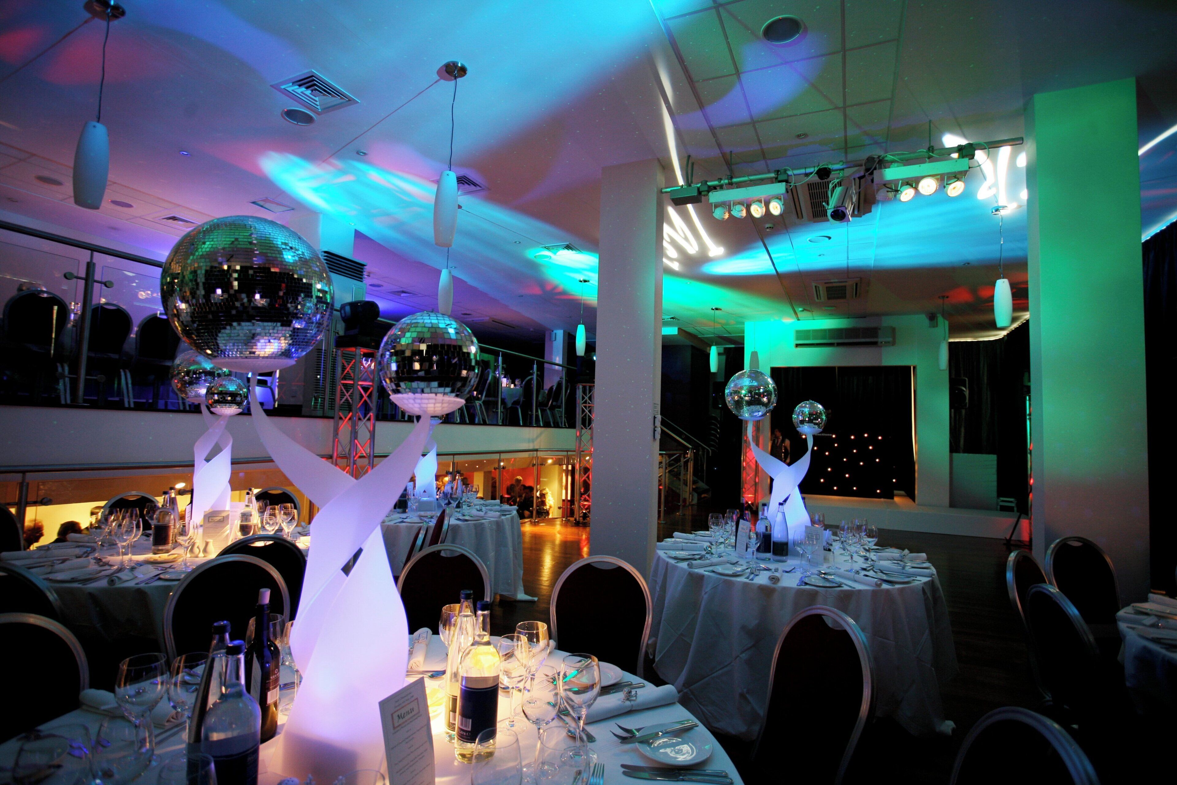 A dining area with tables set for a meal, overhead disco balls, and a stage with lighting equipment.