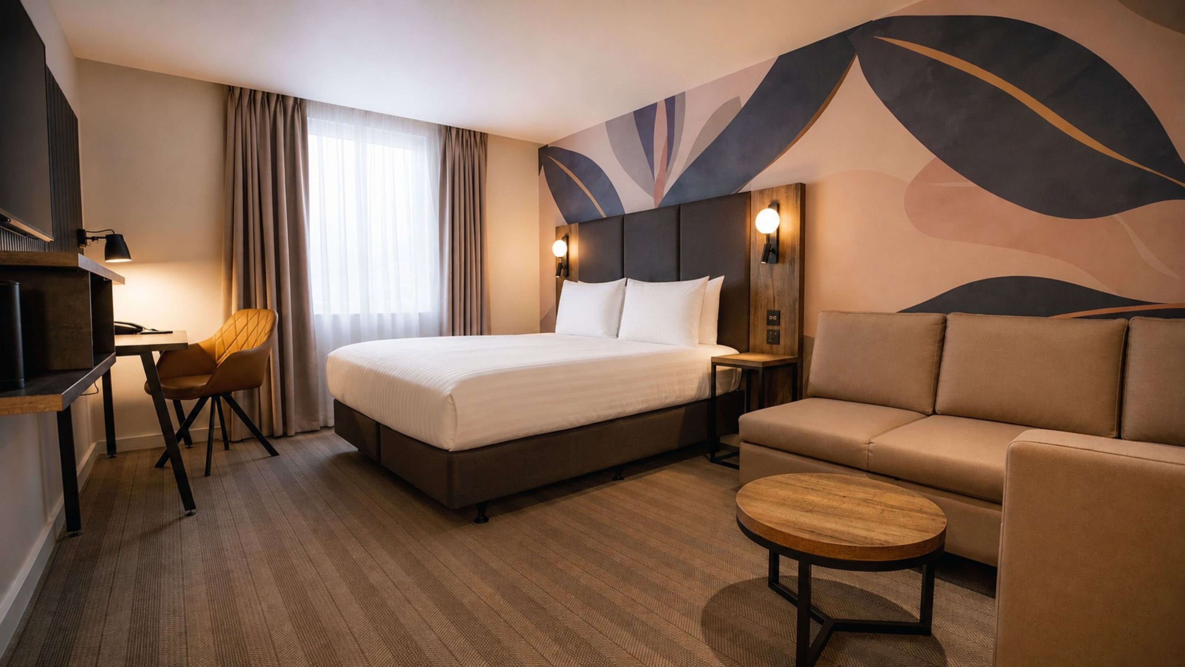 A hotel room with a bed, a sofa, a small table, a desk with a chair, and a TV.