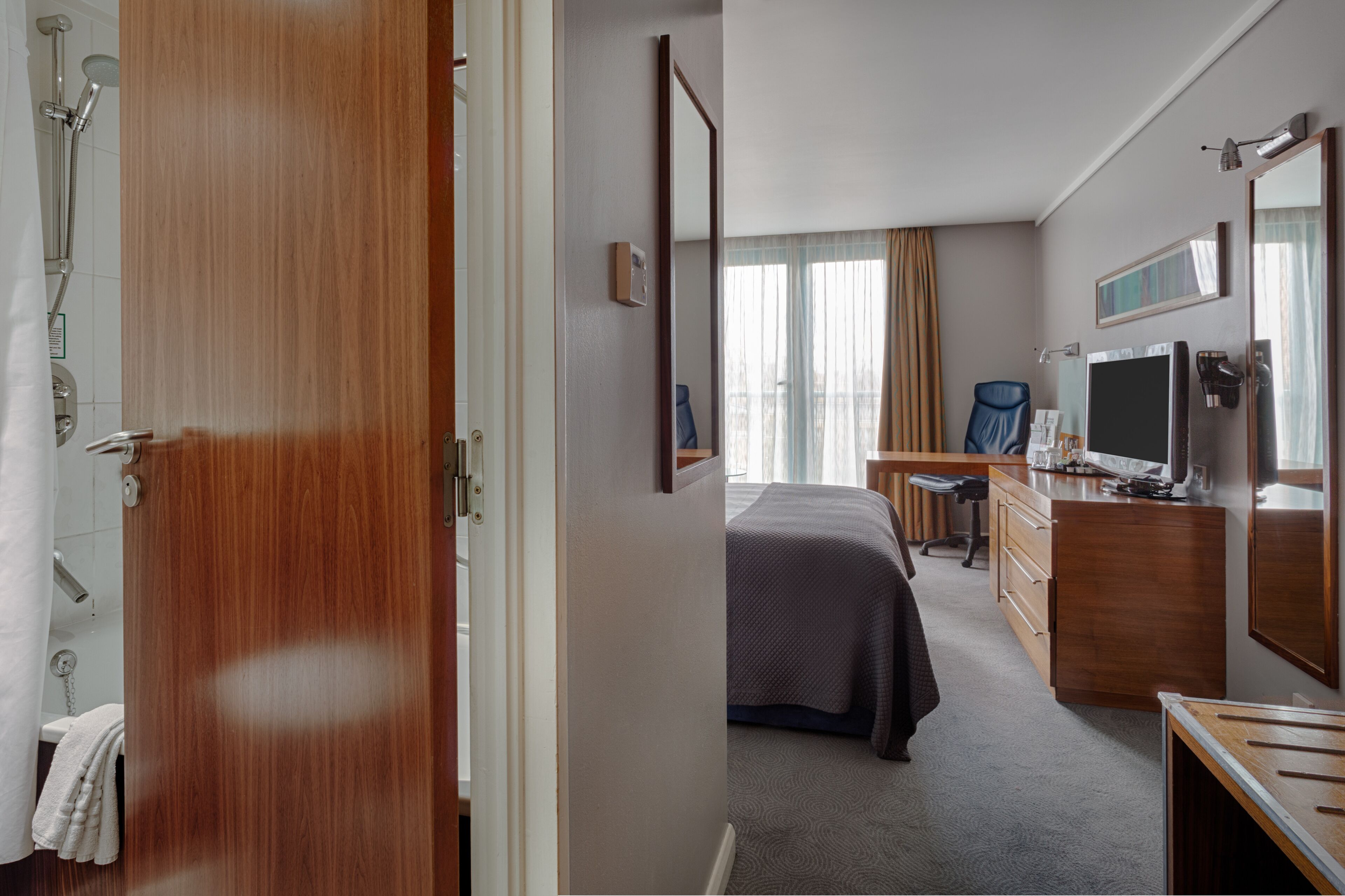 A hotel room with a wooden door, a bed, a desk with a chair, a dresser, a TV, and a large mirror.