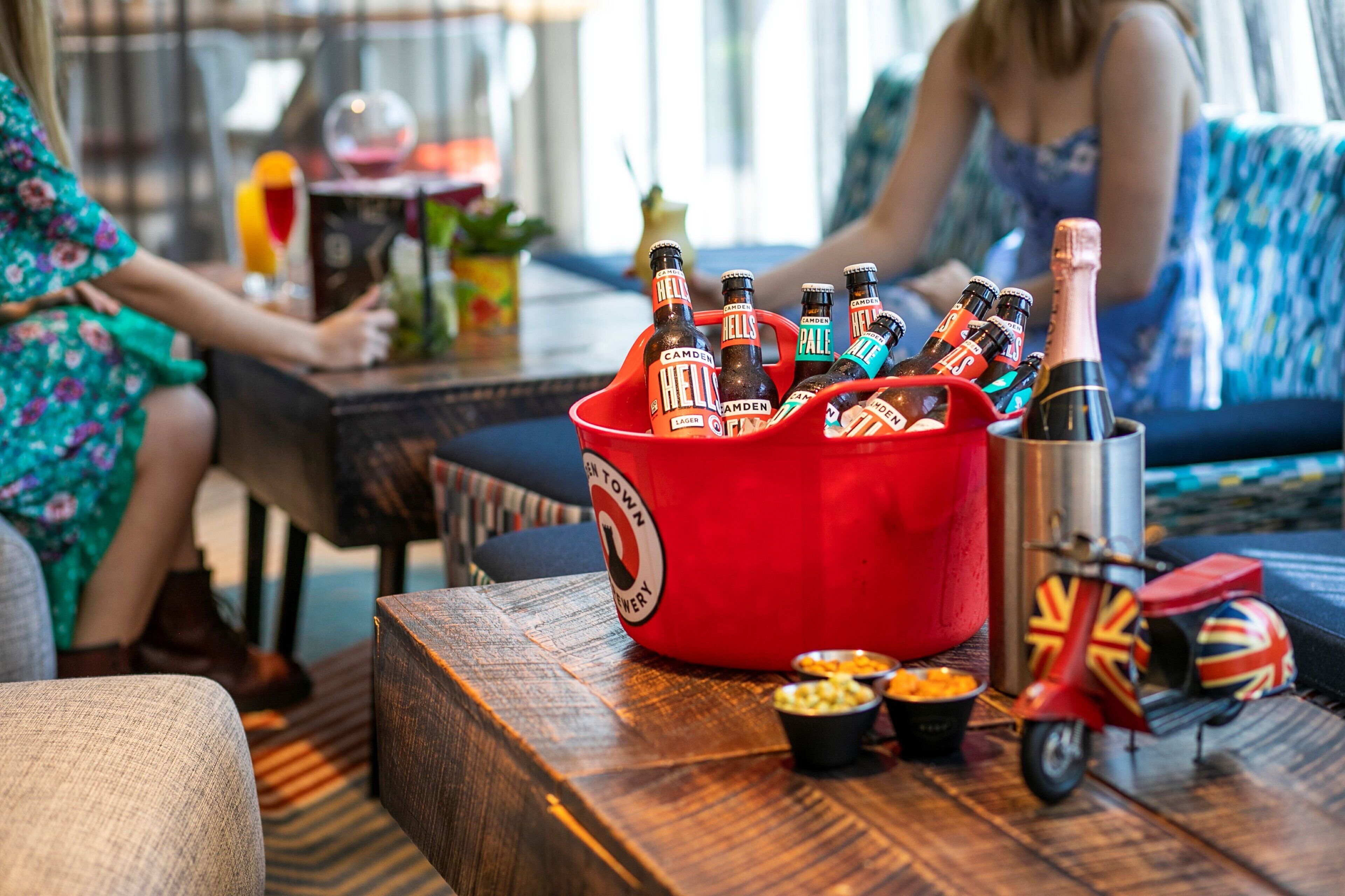 A red bucket filled with beer bottles, a metal cup with a Union Jack flag, and small bowls of snacks on a wooden table in a room with a sofa and a person in the background.