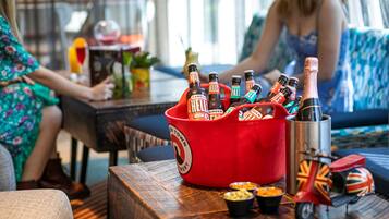A red bucket filled with beer bottles, a metal cup with a Union Jack flag, and small bowls of snacks on a wooden table in a room with a sofa and a person in the background.