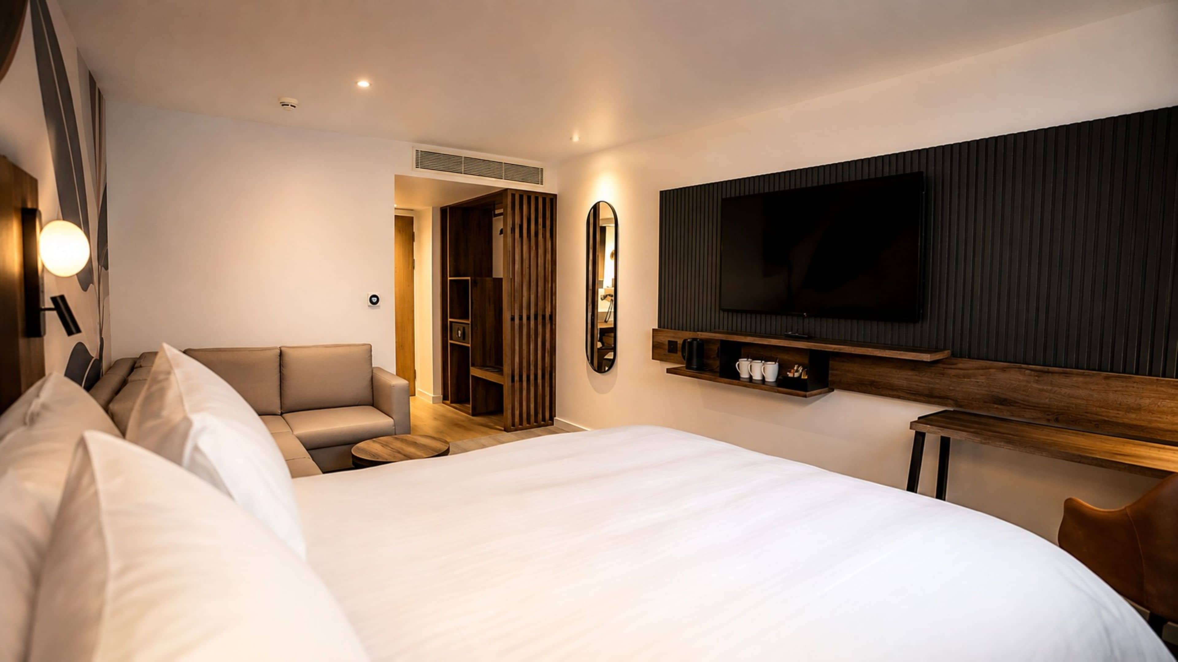 A modern hotel room with a large bed, a television mounted on the wall, a seating area with a sofa, and a wooden cabinet below the TV.