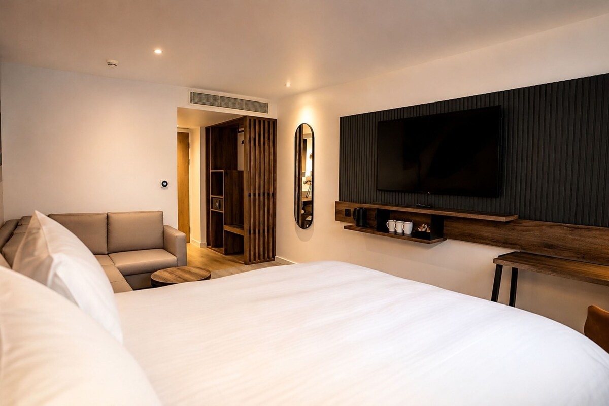 Una habitación de hotel moderna con una cama grande, un televisor montado en la pared, una zona de estar con sofá y un mueble de madera debajo del televisor.