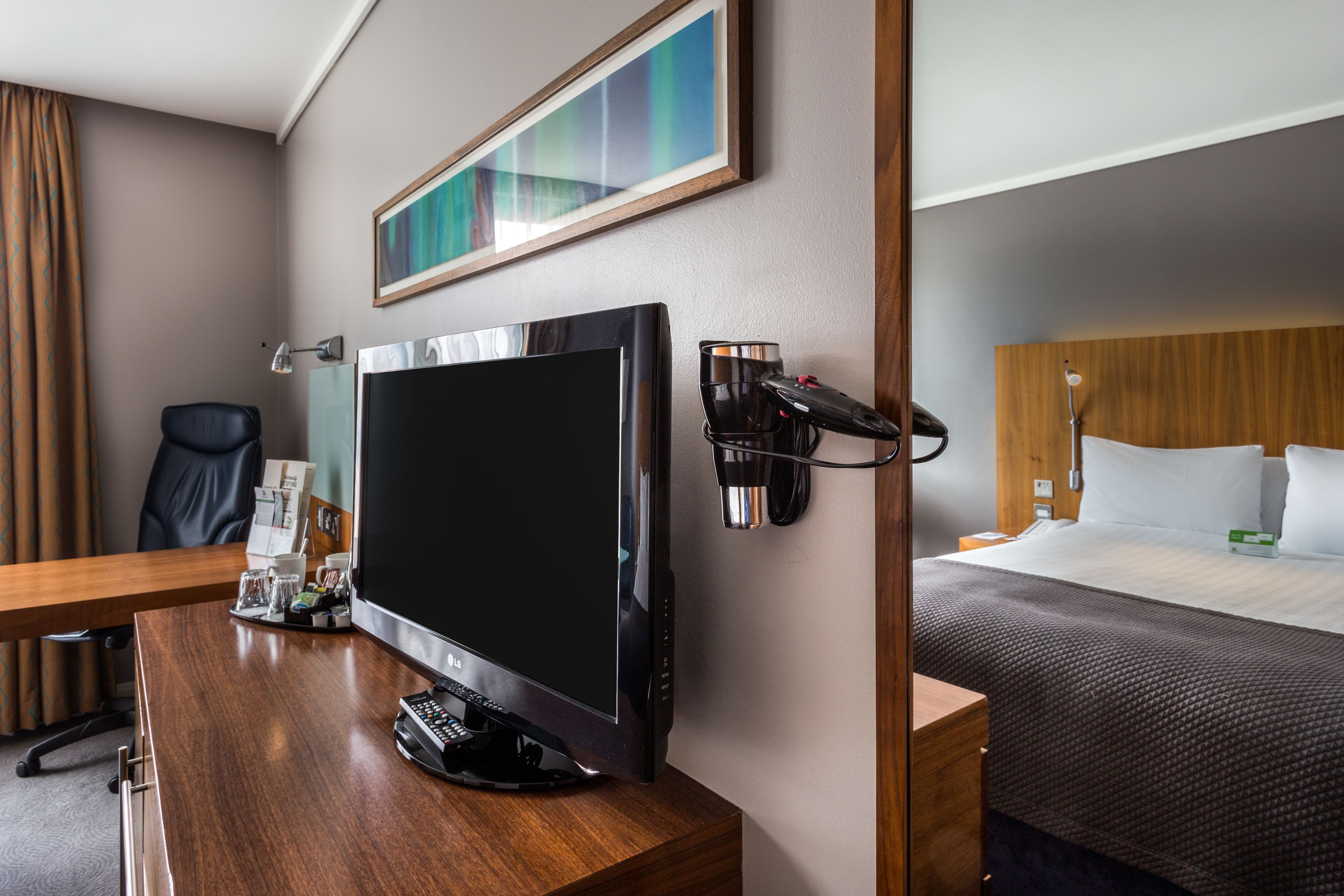 Ein Hotelzimmer mit Holztisch, Flachbildfernseher, Bett mit Holz Kopfteil, Nachttisch und Wandlampe.