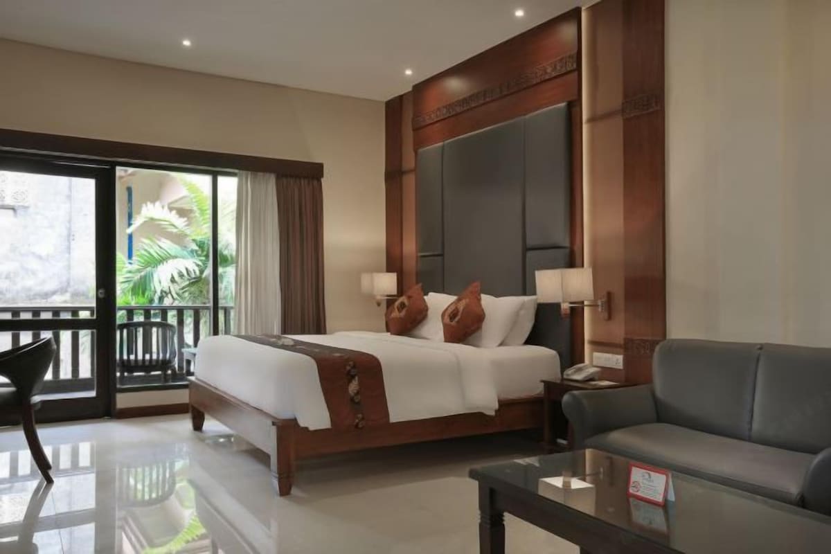 A hotel room with a large bed, a sofa, a glass coffee table, and a balcony with a view of greenery.