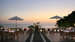 An outdoor dining area with tables and chairs, two umbrellas, and a view of the sea at sunset. - Pelangi Bali Hotel & Spa (Seminyak)