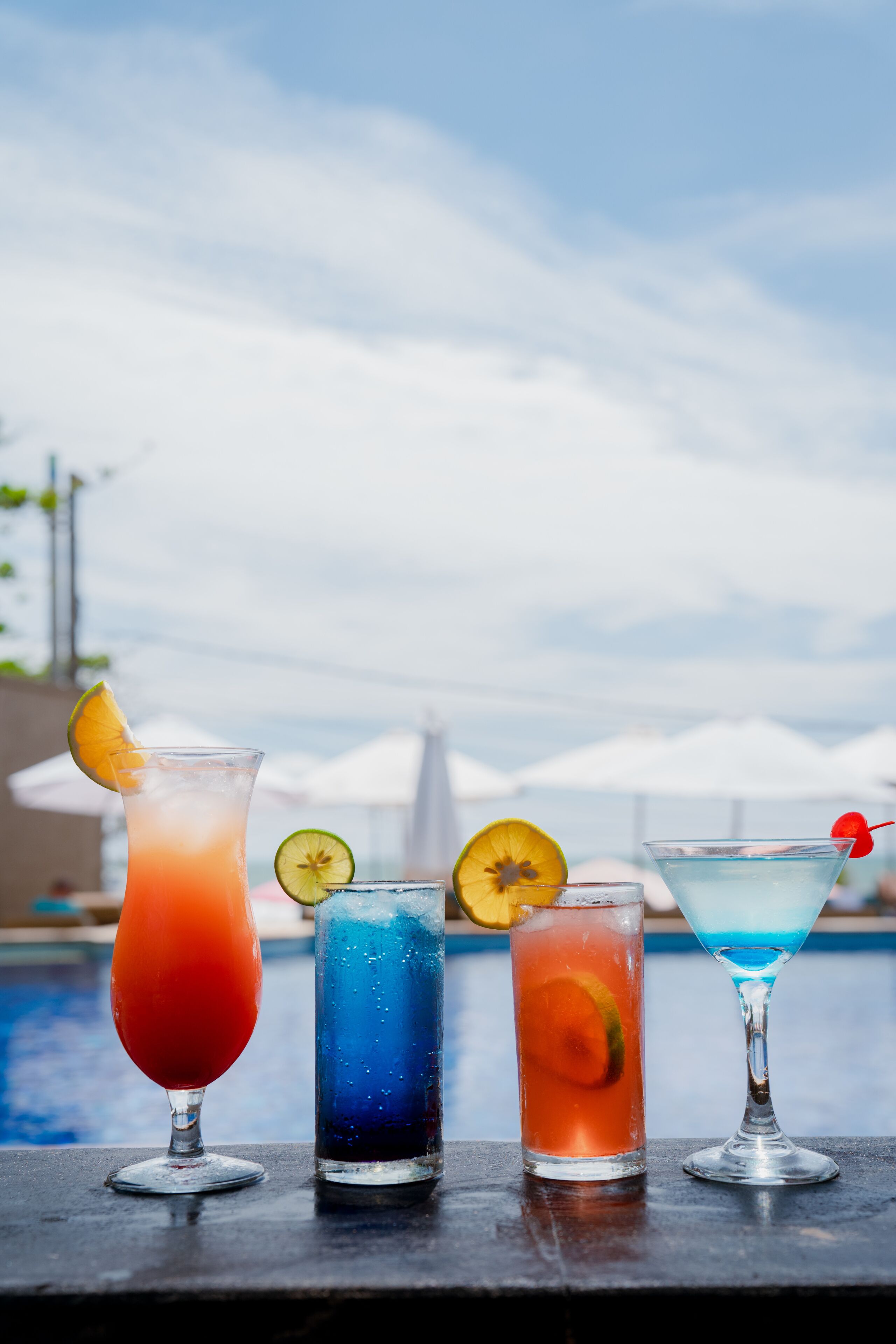 Quatre cocktails, décorés de différents éléments, sont posés sur une table au bord d’une piscine.