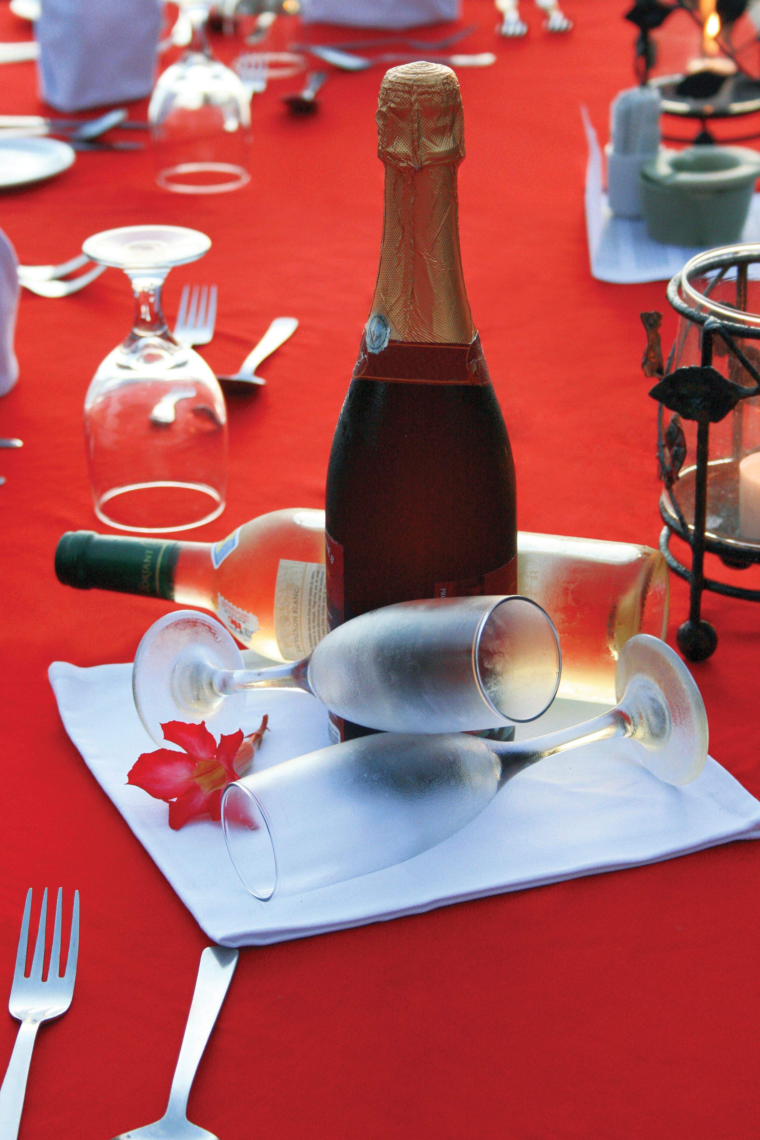 Mesa posta com uma garrafa de champanhe, taças de vinho e talheres sobre toalha de mesa vermelha.