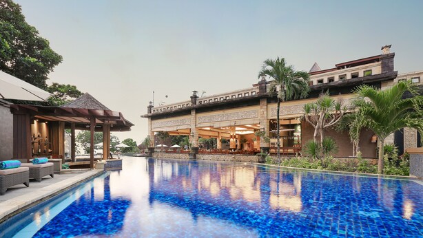 A swimming pool with a tiled surface, a covered seating area with lounge chairs, and a building with a traditional architectural style in the background. - Pelangi Bali Hotel & Spa (Seminyak)