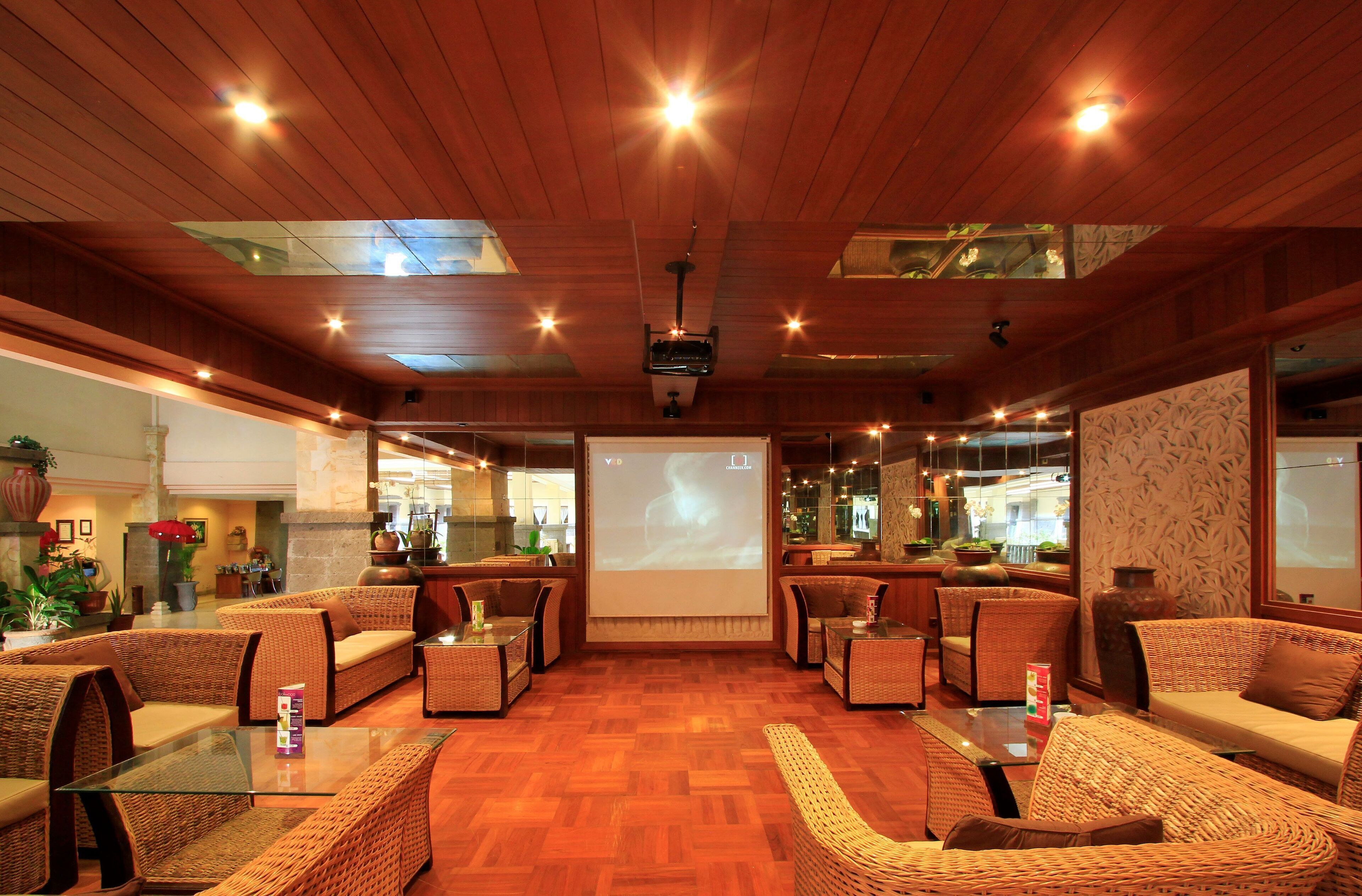 A spacious lobby with wicker furniture, a large screen displaying a projection, and a wooden ceiling with skylights.