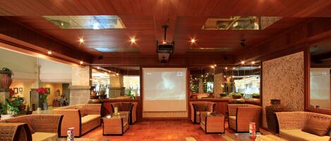 A spacious lobby with wicker furniture, a large screen displaying a projection, and a wooden ceiling with skylights.