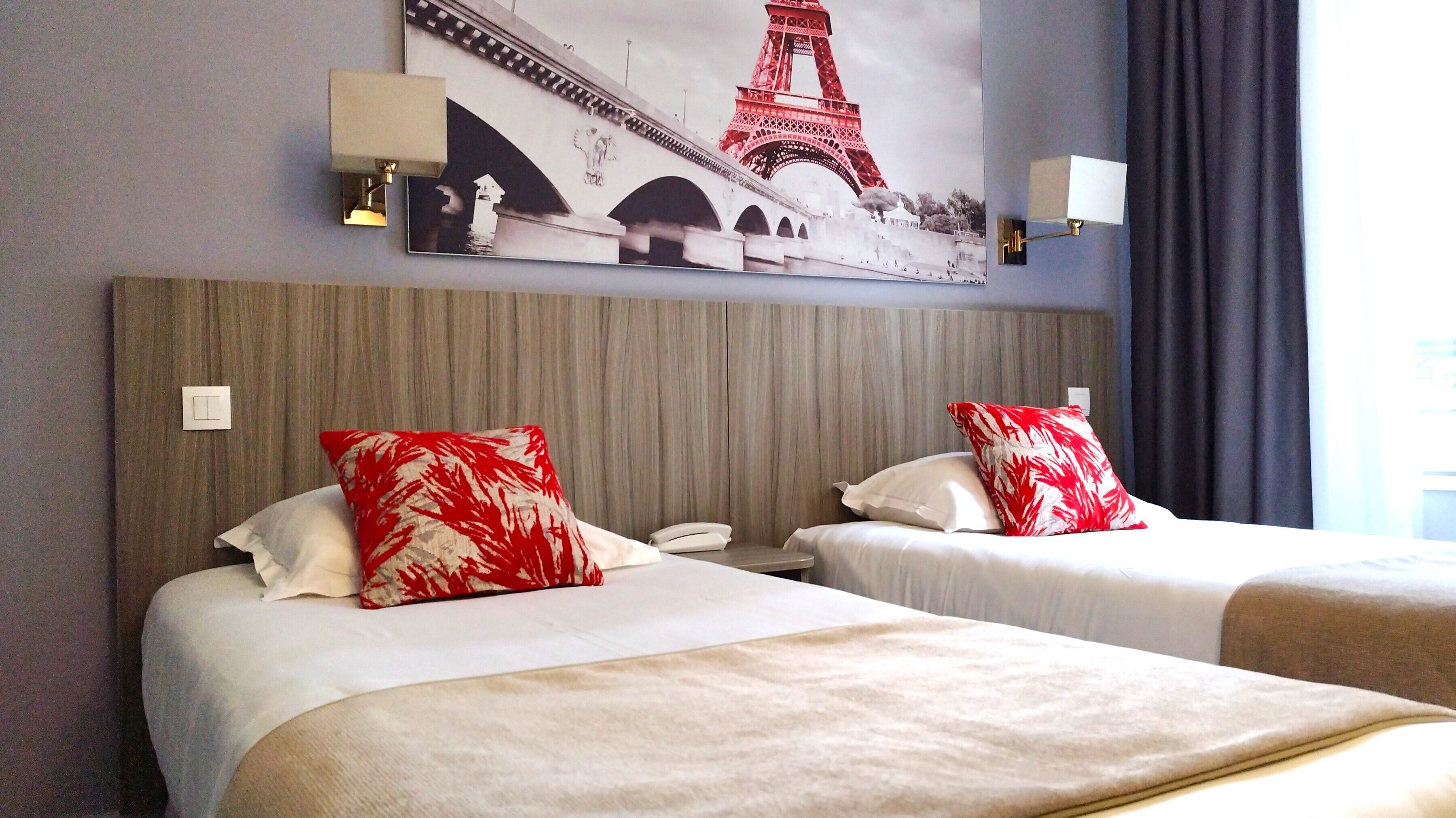 A hotel room with two beds, each with a white bedspread and red and white patterned pillows. There is a large painting of the Eiffel Tower above the beds, and two wall-mounted lamps on either side of the painting.