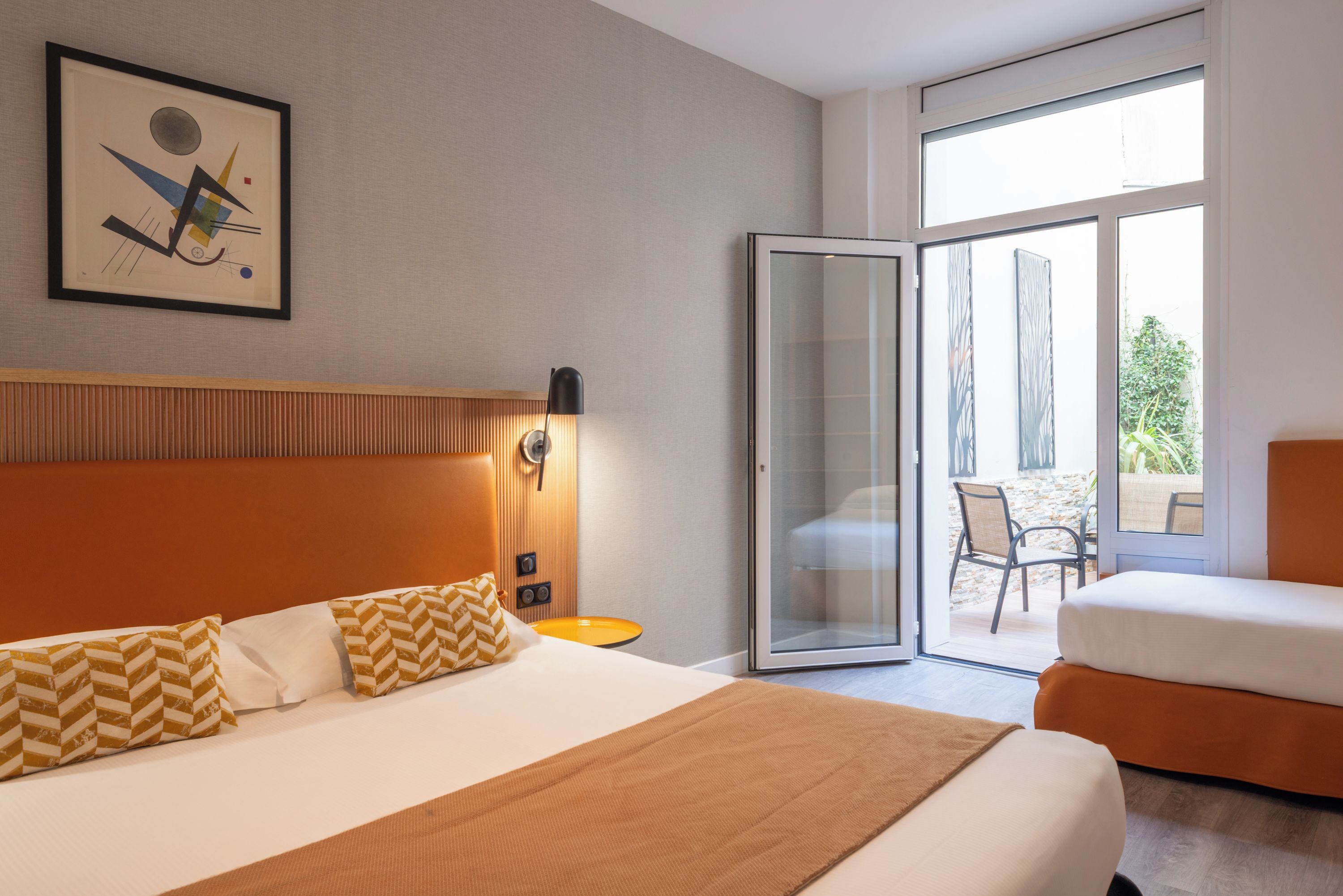 A hotel room with a bed, a chair, a sliding glass door leading to an outdoor area, and a wall-mounted artwork.
