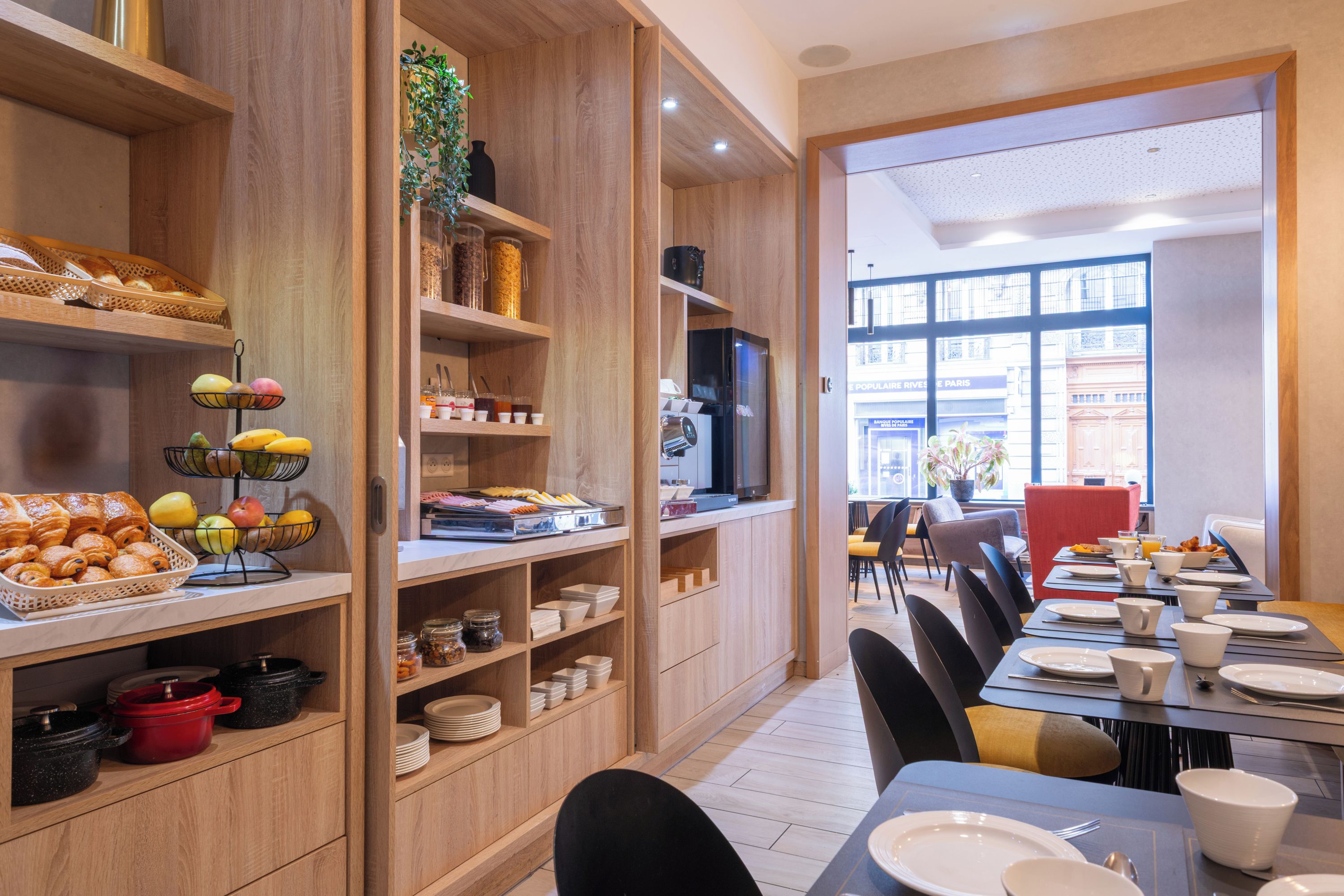 Une salle à manger d’hôtel avec des meubles en bois, un buffet proposant des fruits et des pâtisseries, et une vue sur la rue.