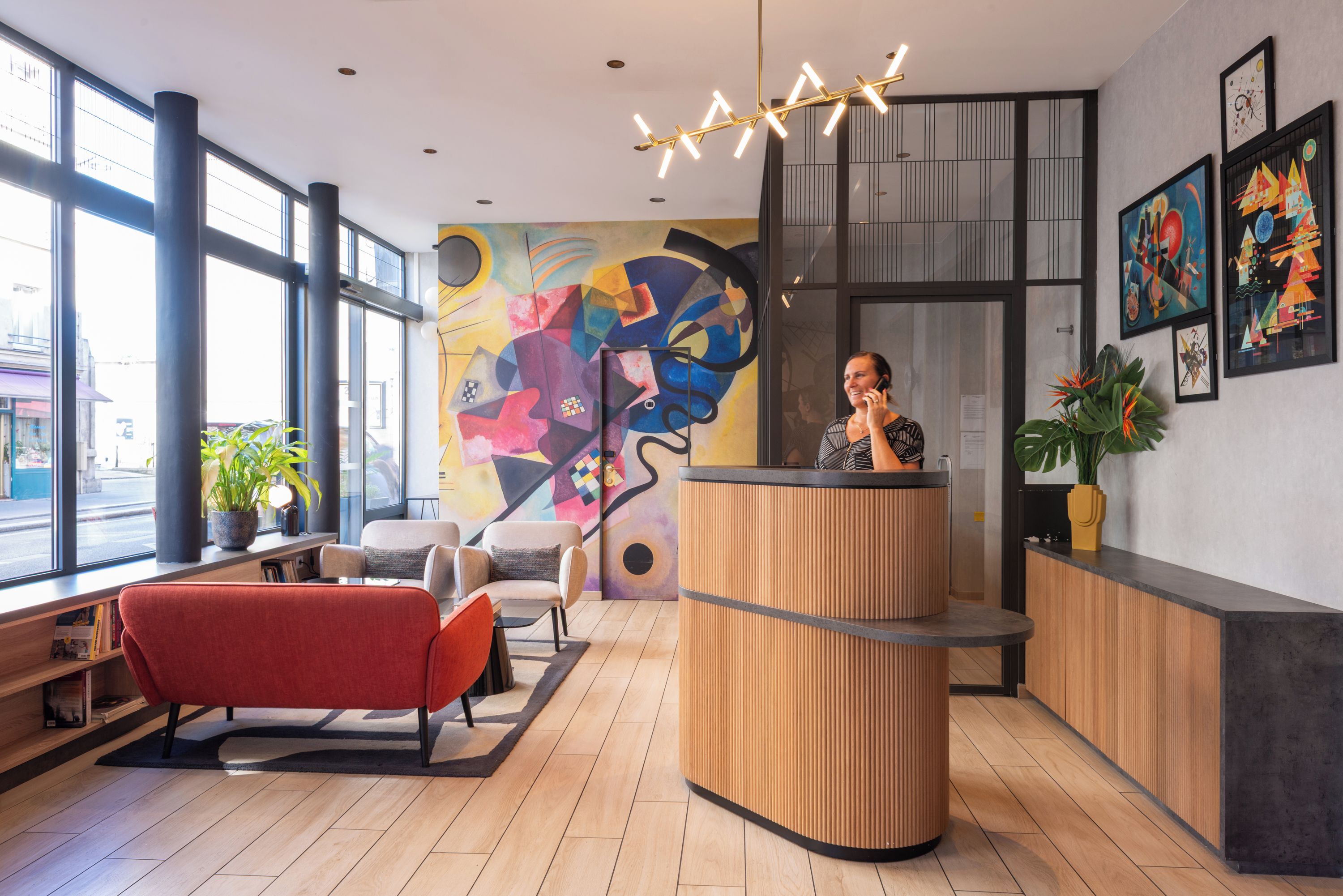 Un espace d’accueil avec un comptoir de réception en bois, un canapé rouge et une fresque murale abstraite et colorée.