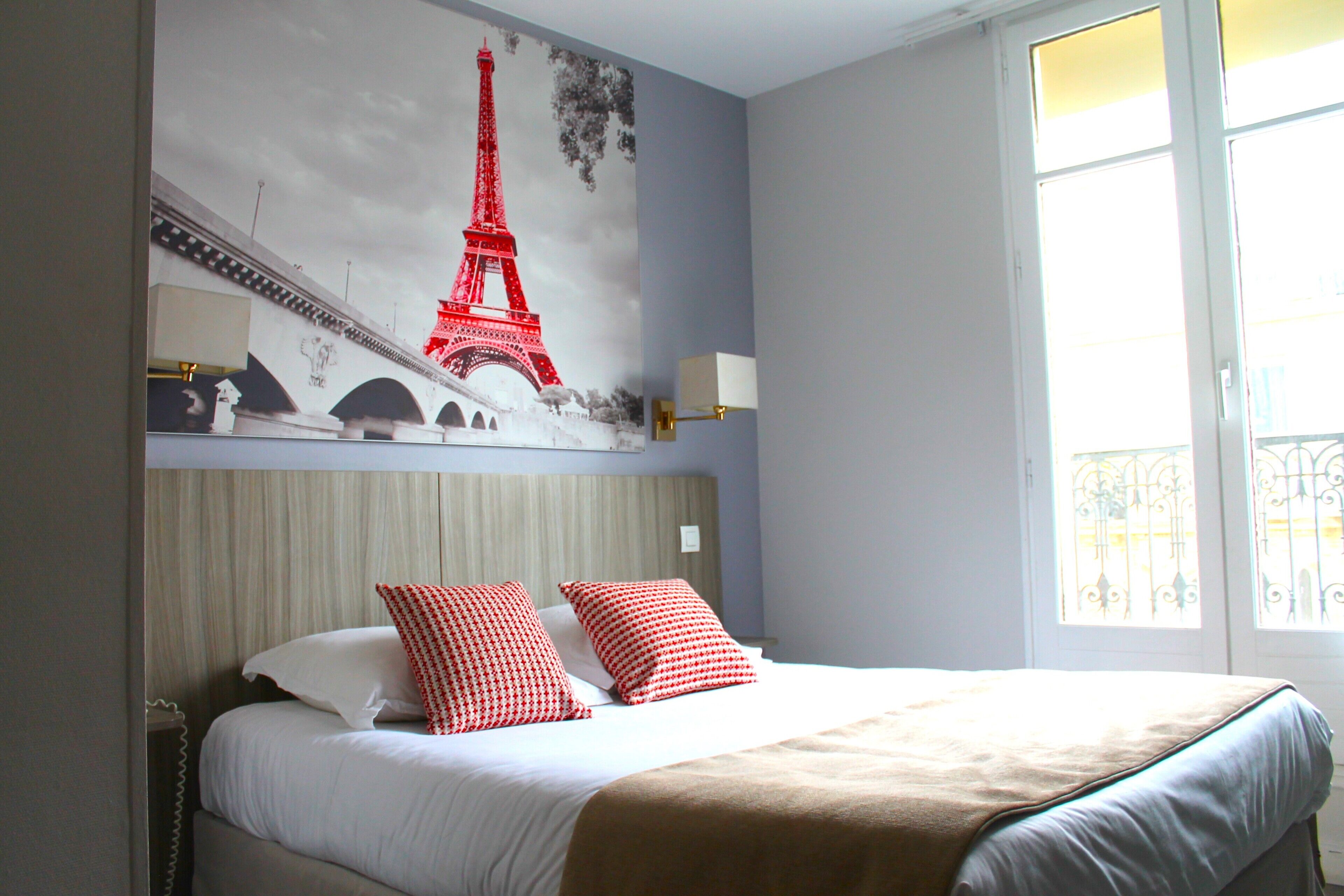 A hotel room with a bed, a large wall mural of the Eiffel Tower, a window with a view, and a bedside lamp.