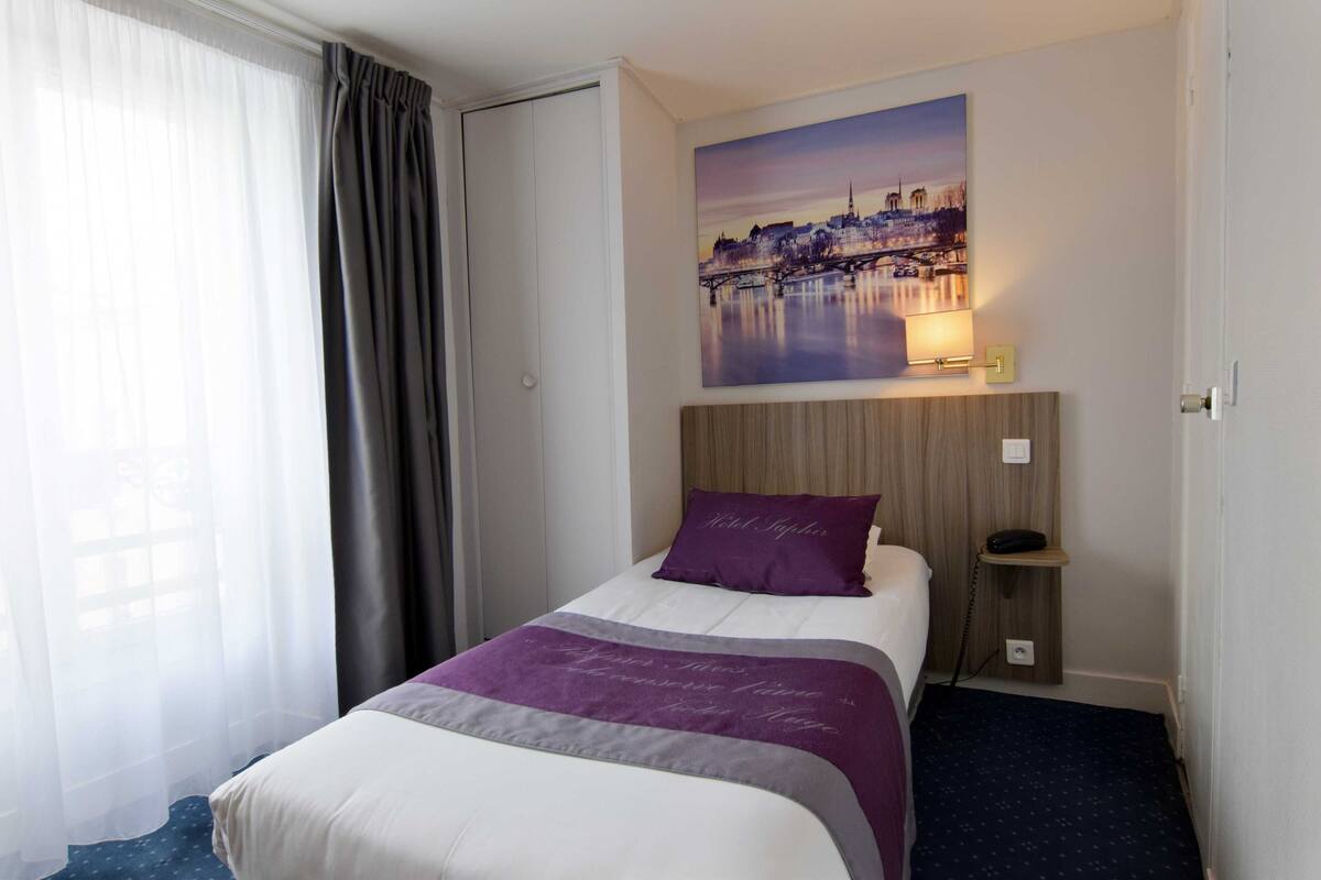Habitación de hotel con cama, mesita de noche, un cuadro de un paisaje urbano y una ventana con cortinas.
