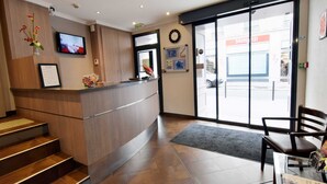 A reception area with a wooden reception desk, a clock, a television, and a view of the outside. - Hôtel Saphir Grenelle (Paris)