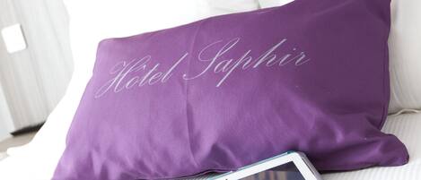 Una cama con cabecera blanca, un almohadón morado con el nombre "Hotel Saphir" bordado, una tableta y un teléfono inteligente sobre la cama.