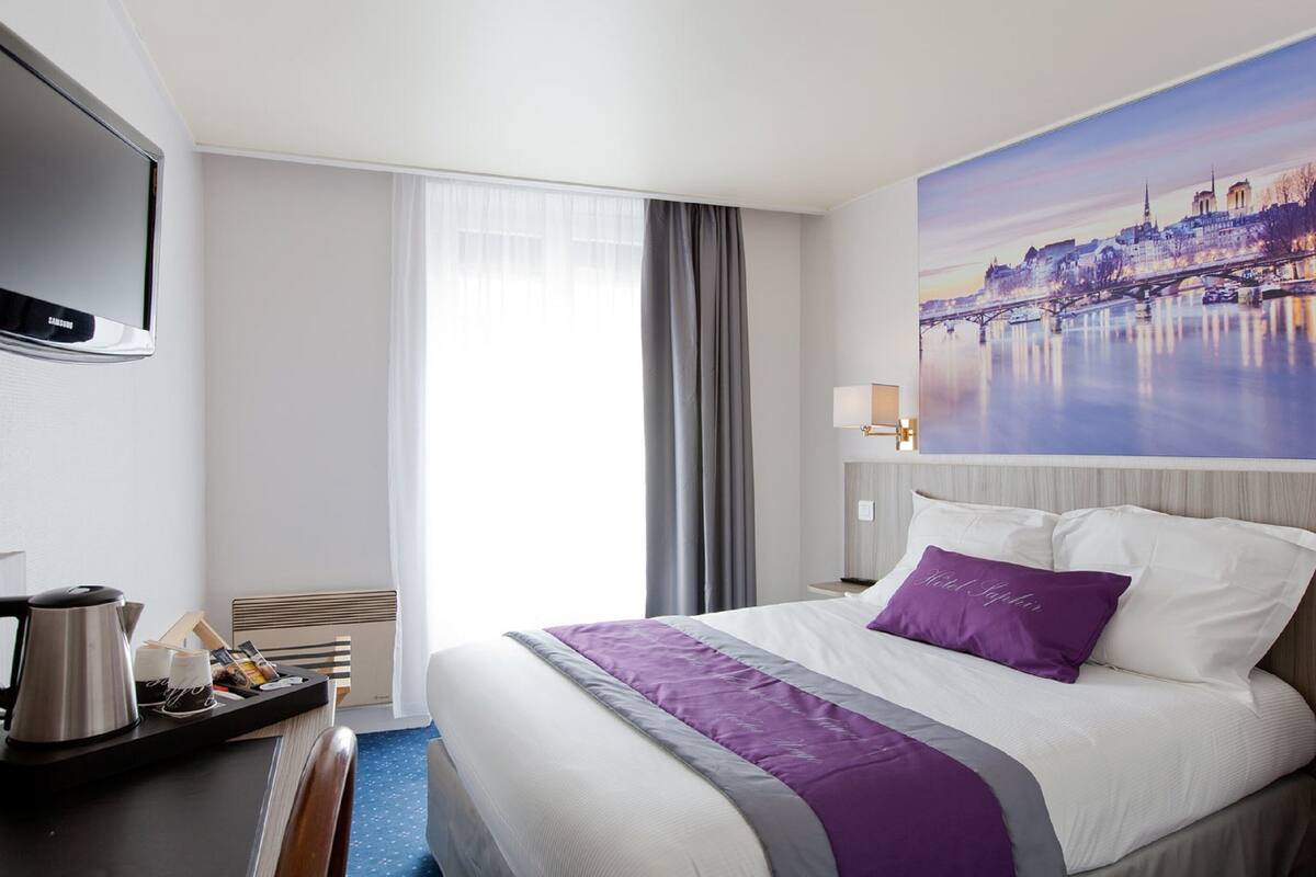 Habitación de hotel con una cama, televisión, un cuadro de un paisaje urbano y una bandeja con cafetera y aperitivos.
