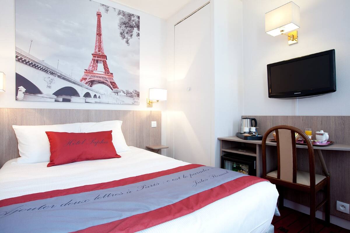 Una habitación de hotel con una cama, un almohadón rojo, un televisor montado en la pared, una cafetera y una silla.