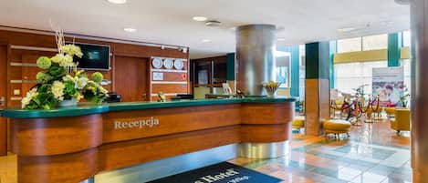 A hotel reception desk with a wooden finish, a digital clock, and a flower arrangement.