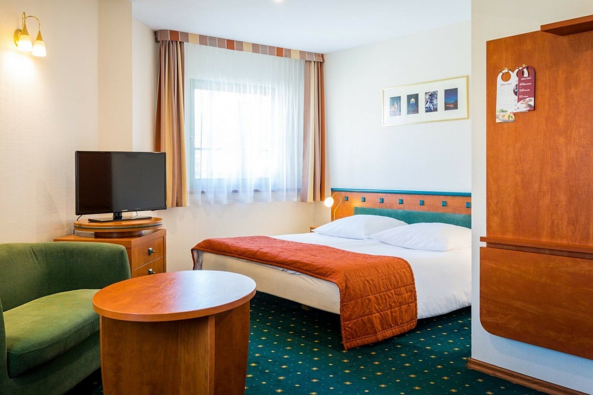 Une chambre d’hôtel avec un lit, une télévision, un fauteuil vert, une table ronde en bois et une armoire.