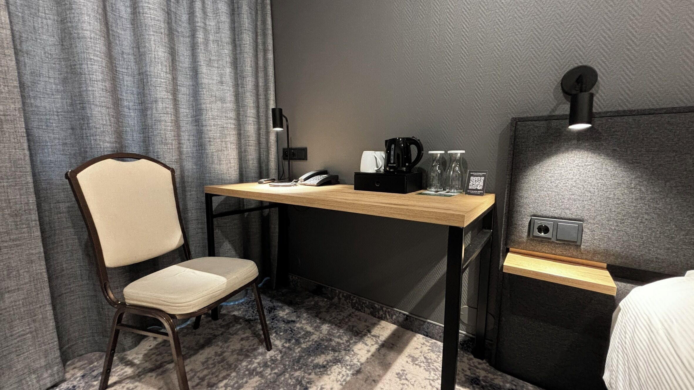 Una habitación de hotel pequeña con una cama individual, un escritorio con teléfono, una cafetera y una mesita de noche con lámpara.