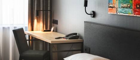 Habitación de hotel con cama, escritorio con teléfono, una silla, lámpara y un cuadro de colores en la pared.