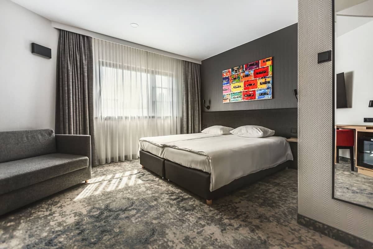 Une chambre d’hôtel moderne comprenant un lit, un canapé, une grande fenêtre avec des rideaux, une œuvre d’art colorée accrochée au mur et un parquet à motifs en chevrons.