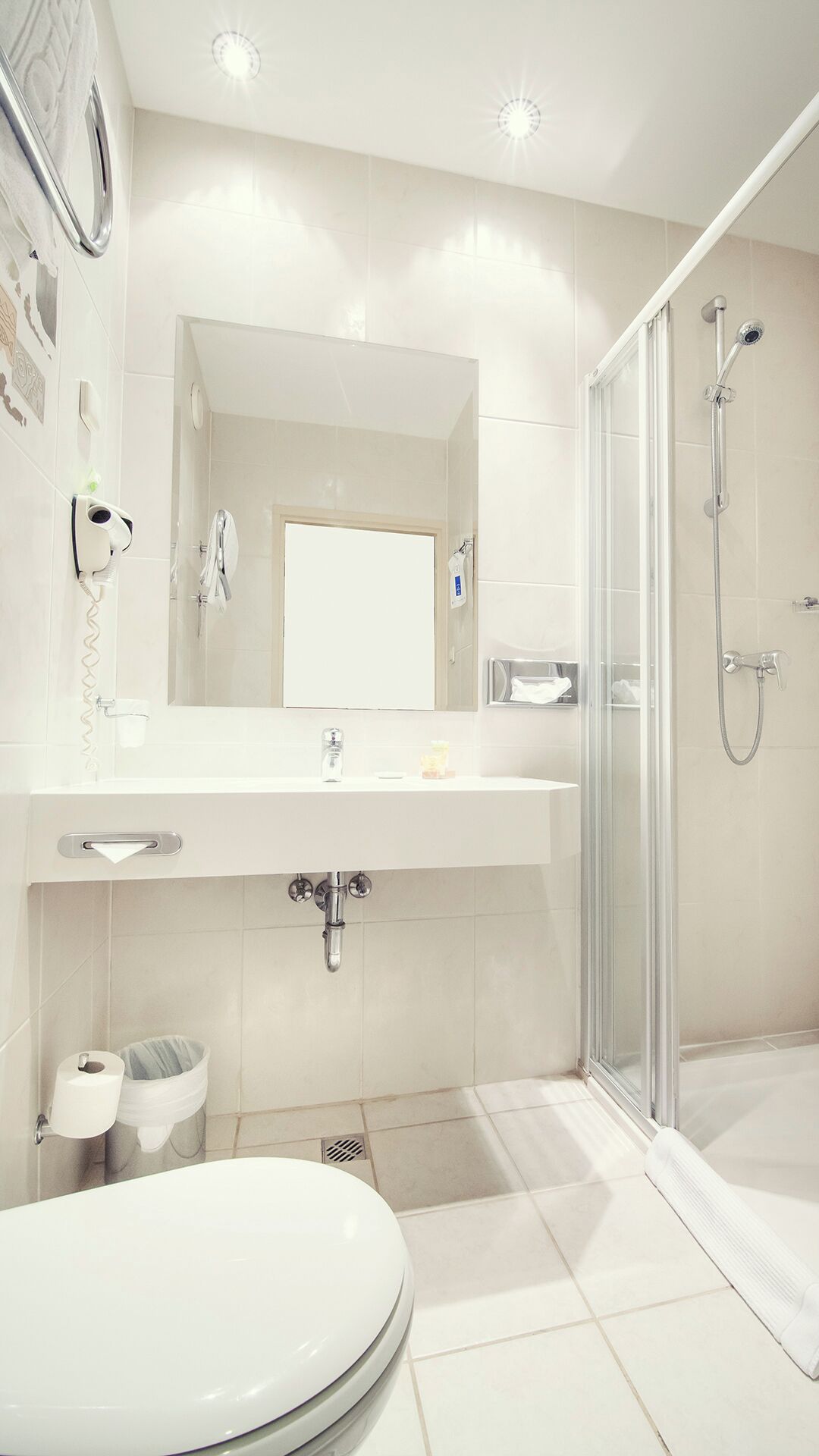 Un baño moderno con cabina de ducha de vidrio, un lavamanos blanco con un espejo encima y un inodoro con dispensador de toallas de papel.