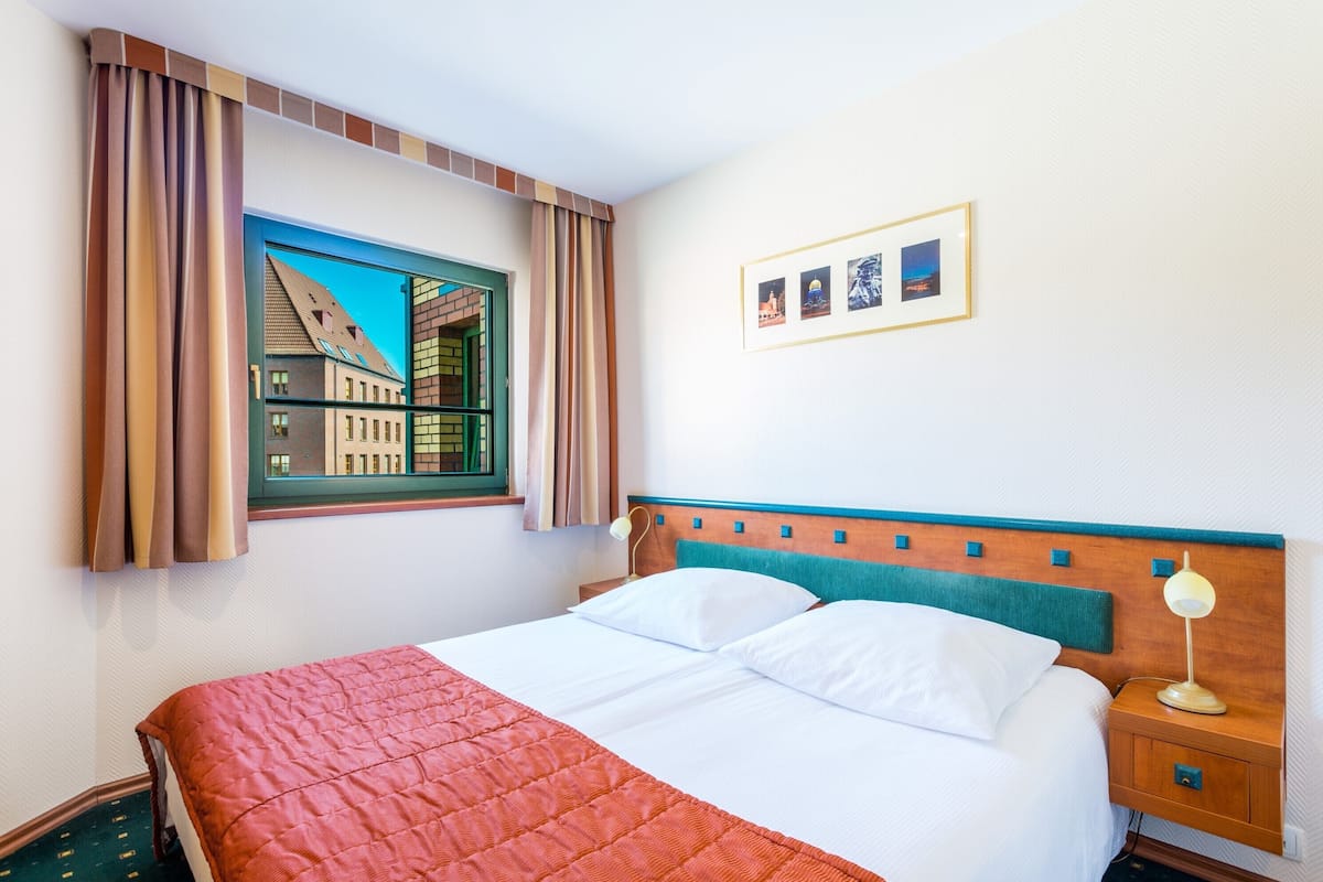 Une chambre d’hôtel comprenant un lit, une table de chevet, une lampe, une fenêtre avec des rideaux et un tableau accroché au mur.