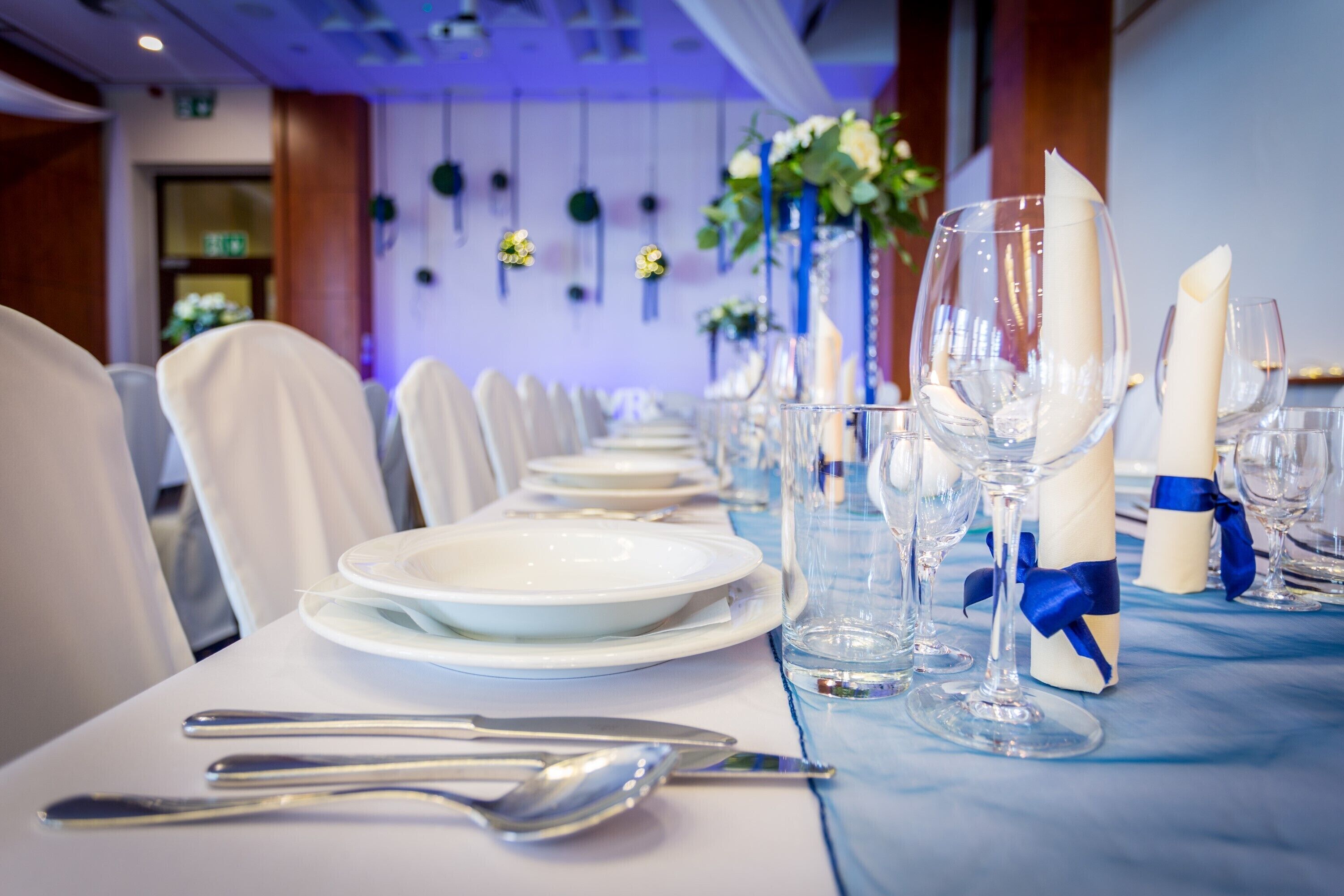 Una larga mesa de comedor dispuesta con platos, cubiertos y vasos, con mantel azul y servilletas blancas atadas con cintas azules.