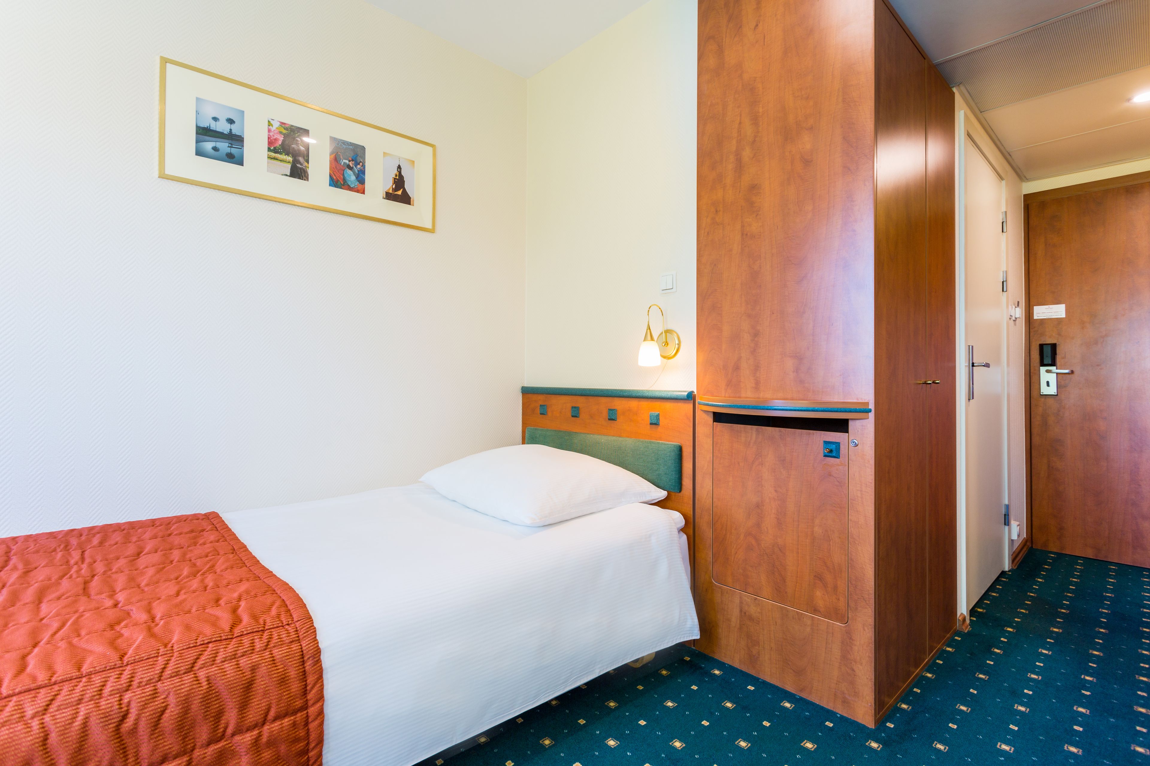 Quarto de hotel com uma cama, um guarda-roupa de madeira, uma fotografia emoldurada na parede e uma mesa pequena com uma luminária.