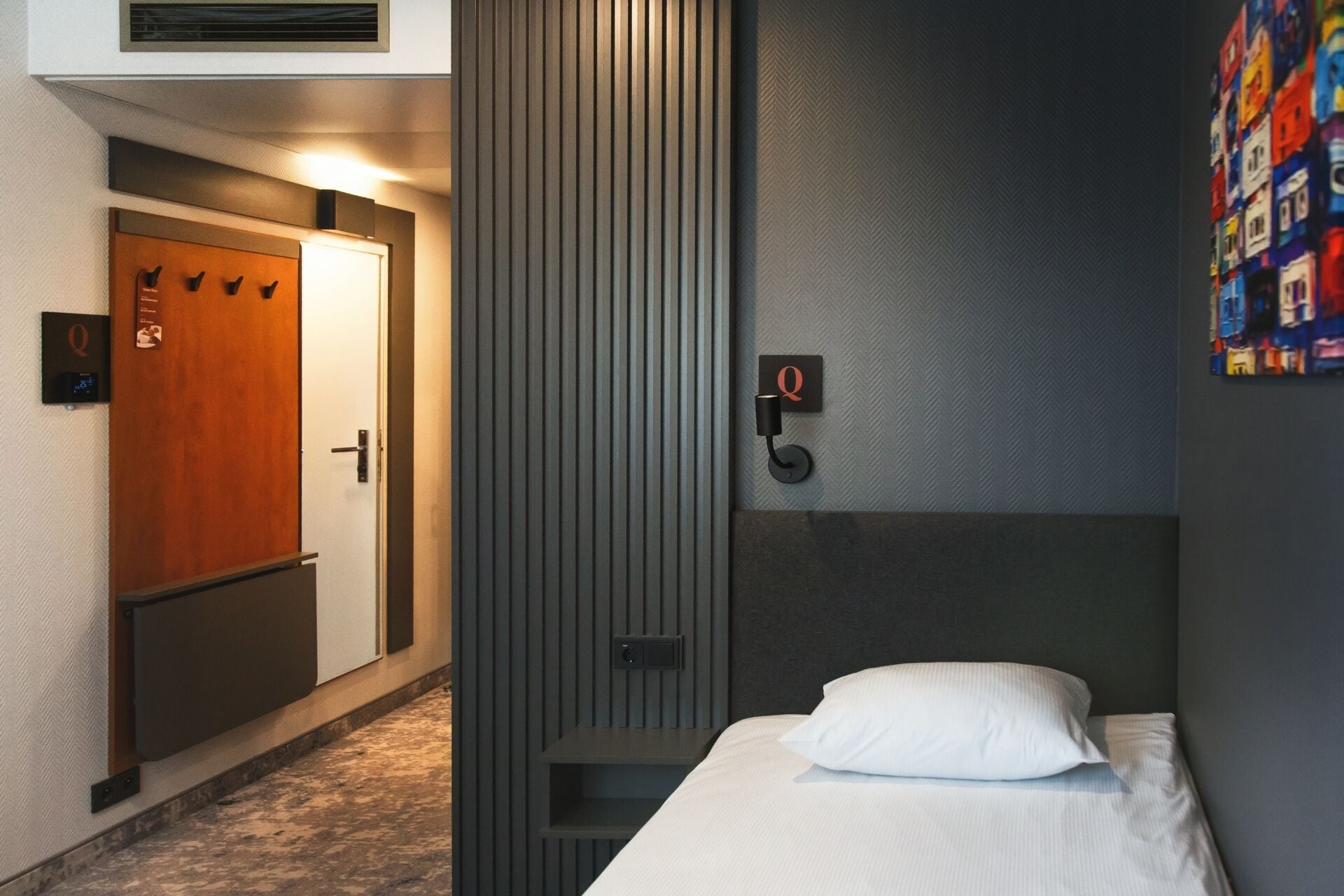 Ett hotellrum med en säng, en trädörr, en vertikal panel och en väggmonterad lampa.