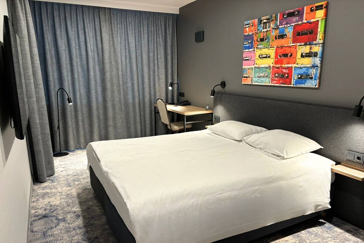 Une chambre d’hôtel avec un grand lit, un bureau, une chaise, une lampe de bureau, un téléviseur fixé au mur et une œuvre d’art colorée accrochée au mur.