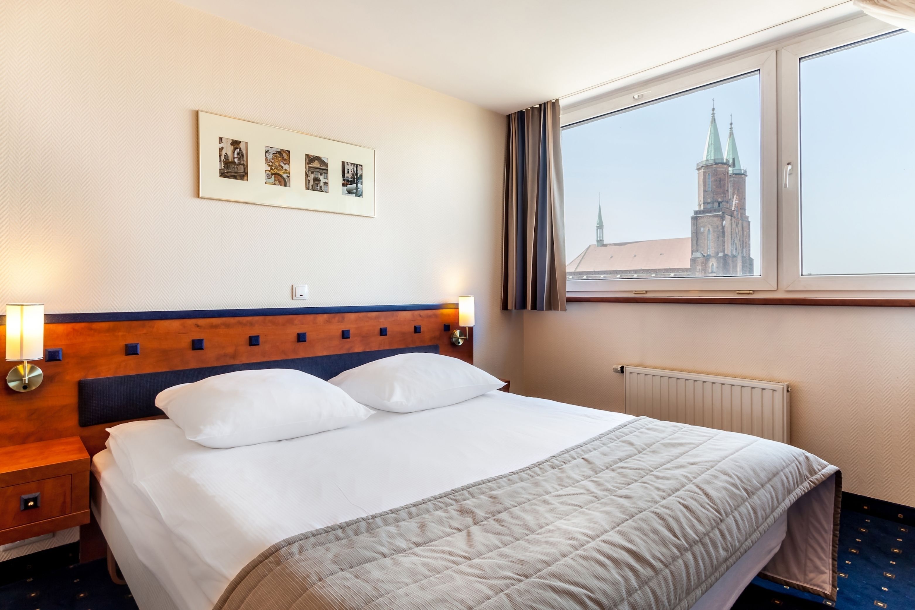 A hotel room with a large bed, a headboard with a wooden panel, a framed picture on the wall, a window with a view of a building, and a nightstand with a lamp.