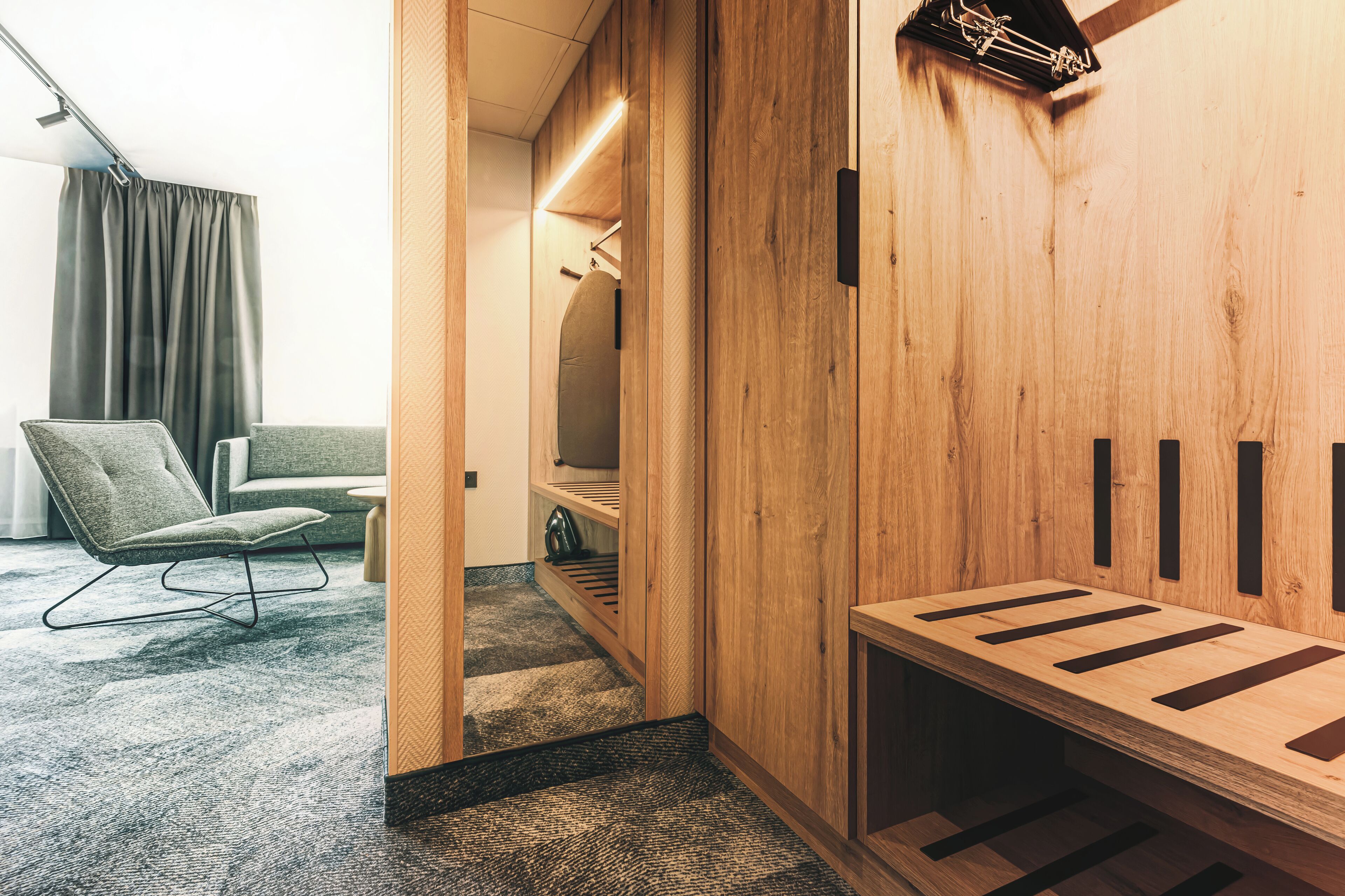 Habitación de hotel moderna con un revestimiento de madera, una zona de estar con una silla y un sofá, y un banco empotrado con espacios de almacenamiento.