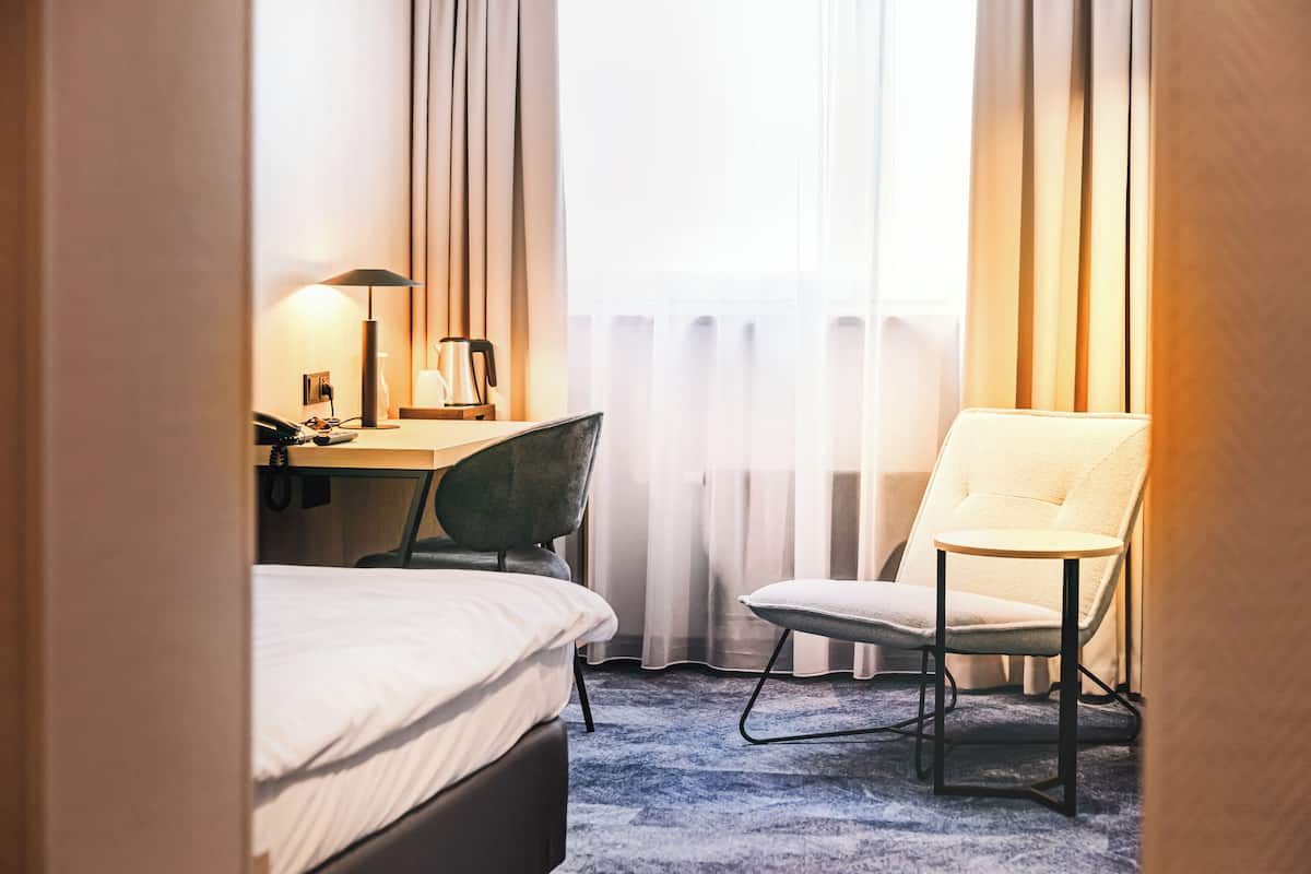 A hotel room with a bed, a desk with a lamp, a chair, and a view of the outside through sheer curtains.